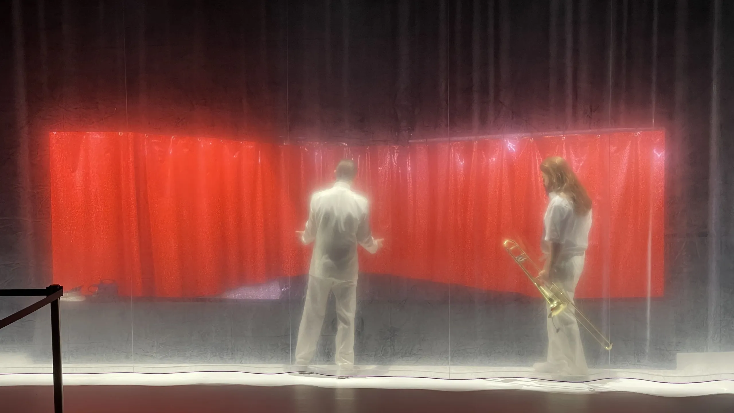 Prøvebilder fra scenen på forestillingen Kallocain - vi ser to skuespillere i hvite klær bak et gjennomsiktig forheng i plast. De står foran noe som er ut som et rødt forheng, og den ene holder en trombone i hånden.