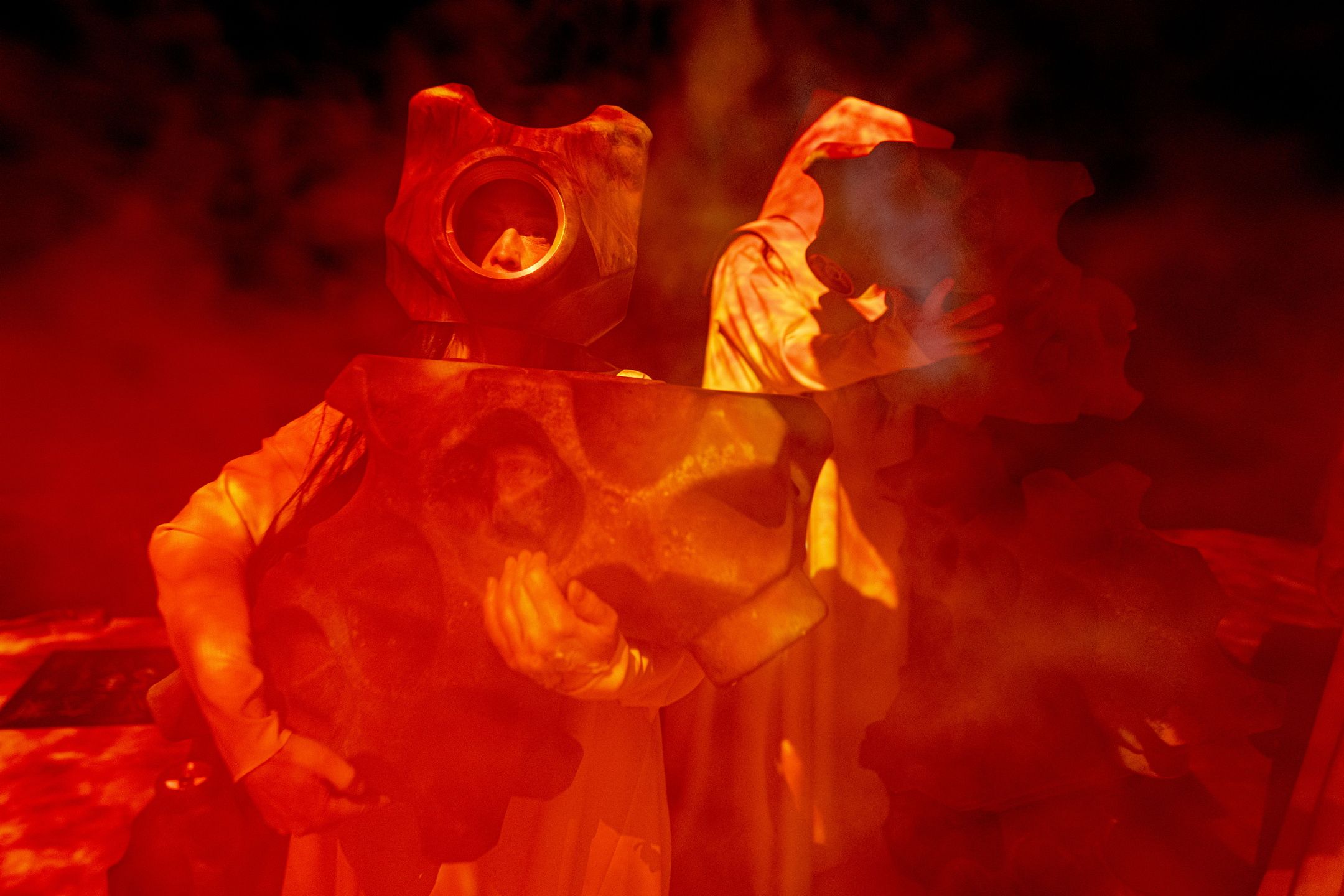 To personer i rødt lys og røyk holder store tannhjullignende masker foran kroppen