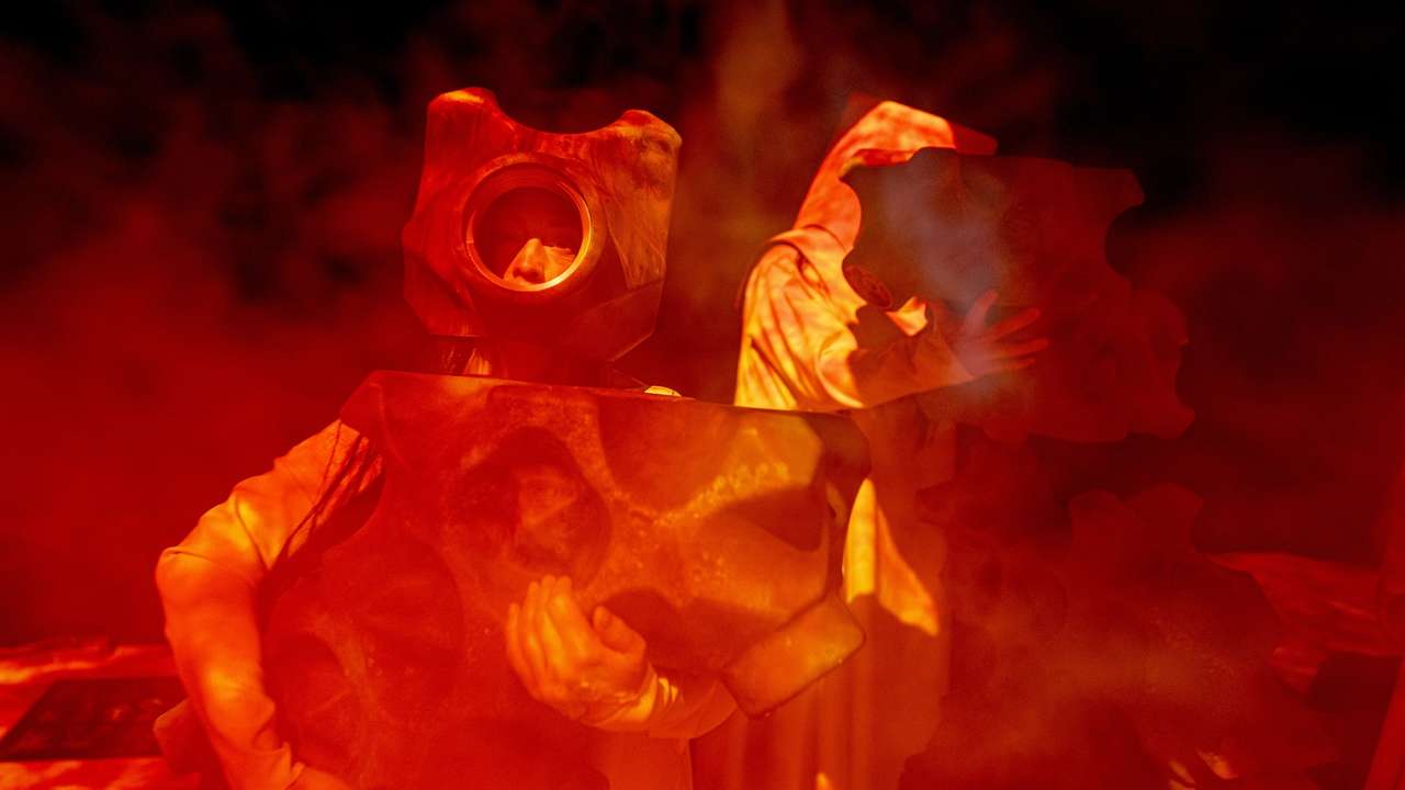 To personer i rødt lys og røyk holder store tannhjullignende masker foran kroppen