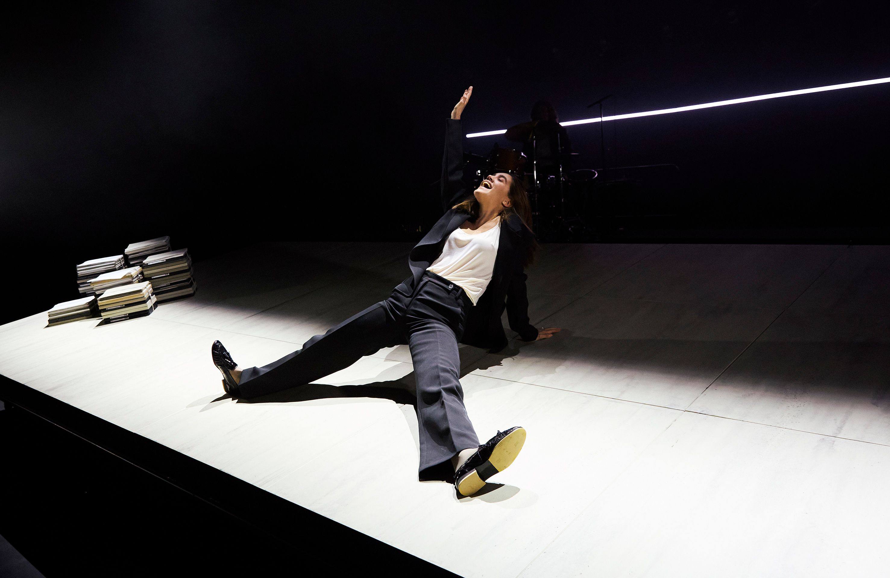 En kvinne i svart dress og hvit topp ligger på gulvet med en arm løftet, bak henne er en trommeslager.