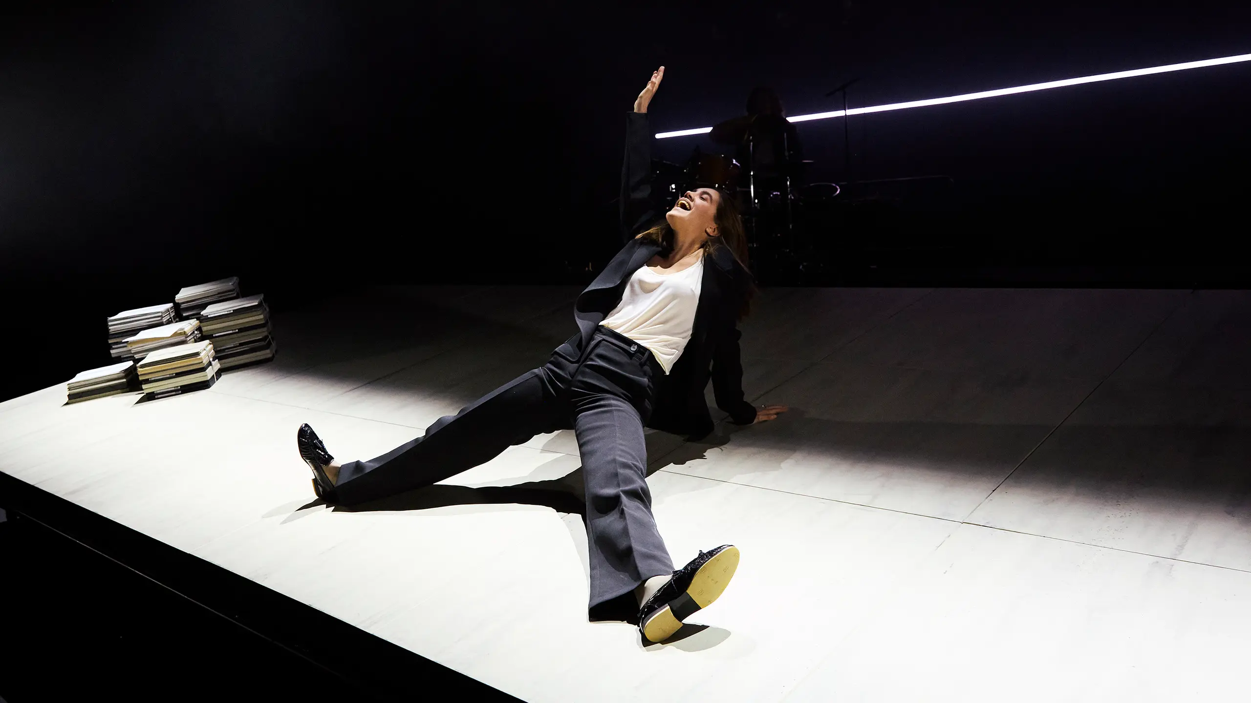 En kvinne i svart dress og hvit topp ligger på gulvet med en arm løftet, bak henne er en trommeslager.