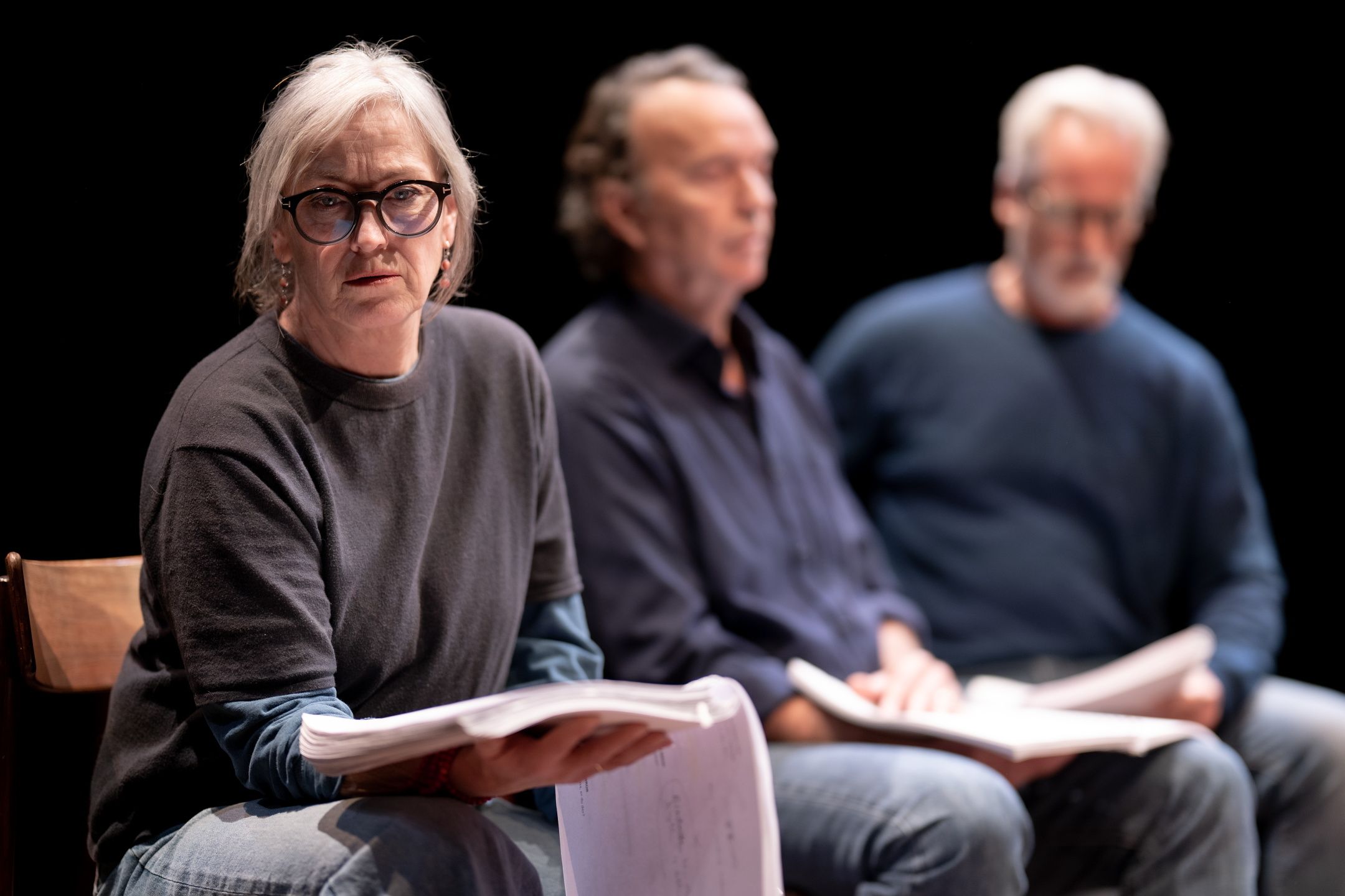 Kvinne som sitter vendt mot kamera med manus. To menn sitter i bakgrunnen