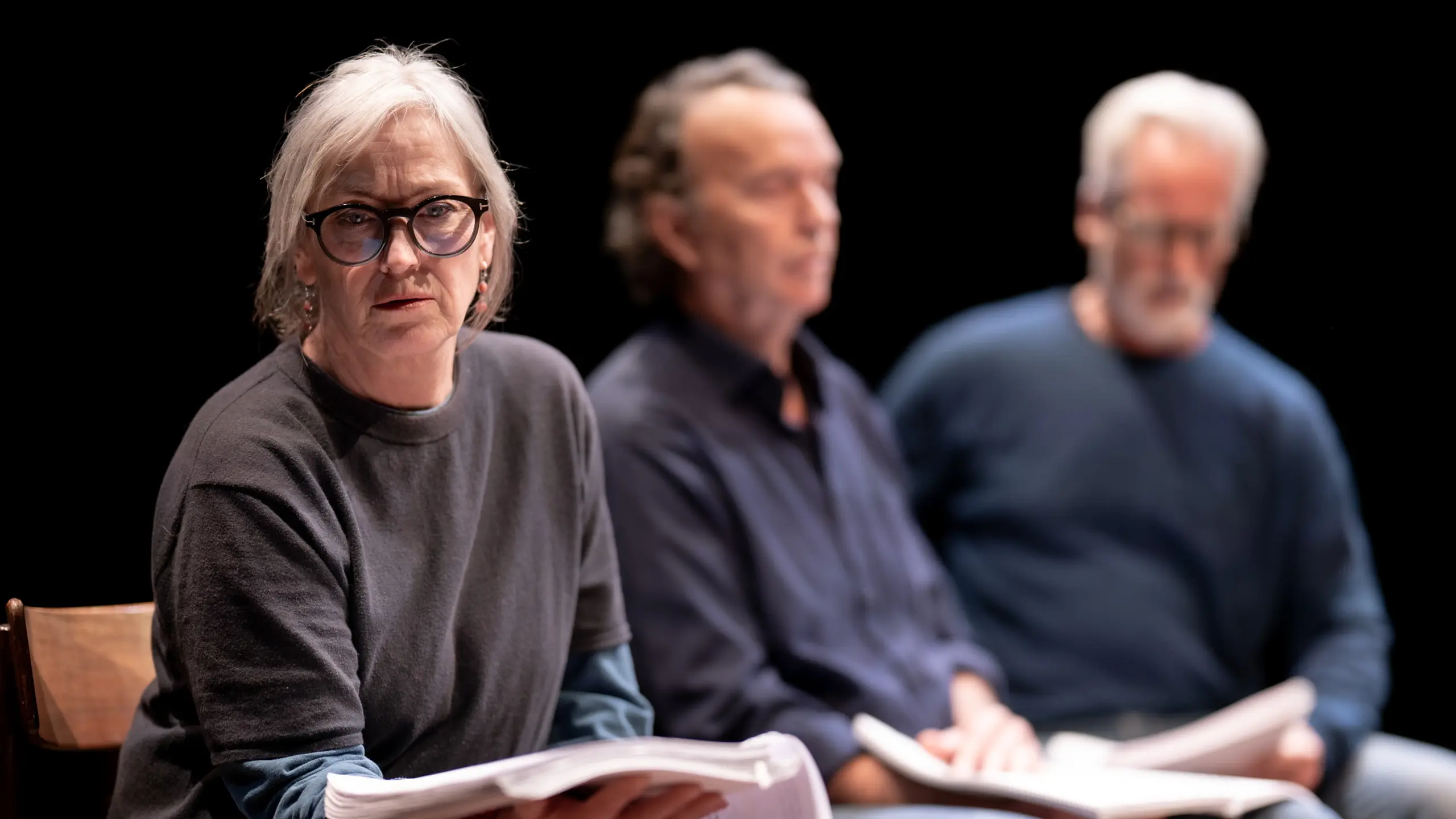 Kvinne som sitter vendt mot kamera med manus. To menn sitter i bakgrunnen