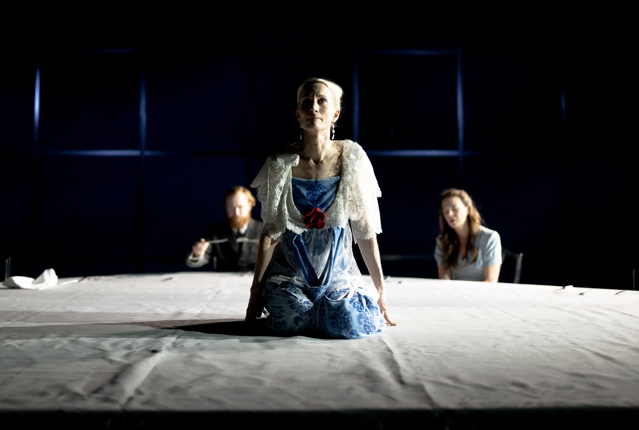 Kvinne i blå og hvit kjole sitter på knærne på et bord med to personer i bakgrunnen.