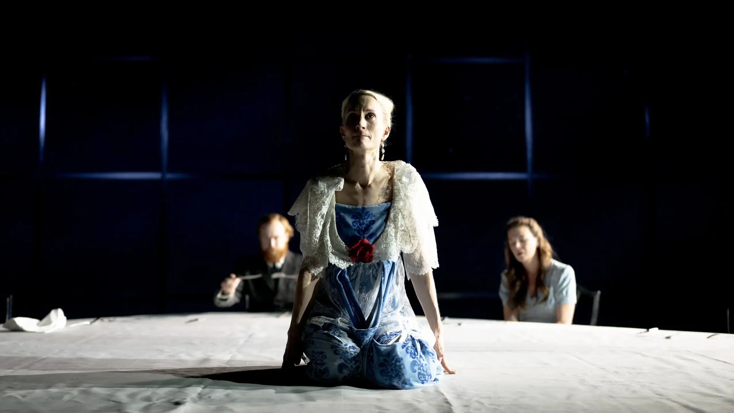 Kvinne i blå og hvit kjole sitter på knærne på et bord med to personer i bakgrunnen.