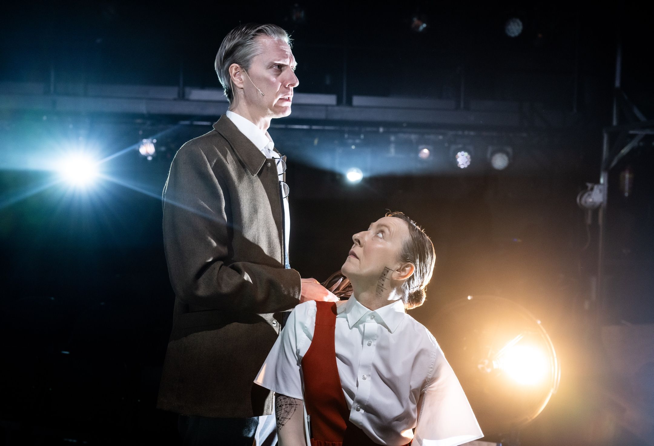 To skuespillere i scenelys: en mann står og ser bestemt fremover mens en kvinne sitter foran ham og ser opp mot ham. Begge har alvorlige ansiktsuttrykk og påtegnet mønster på halsen. Lyset bak dem fremhever silhuetter og skaper en teatralsk stemning.