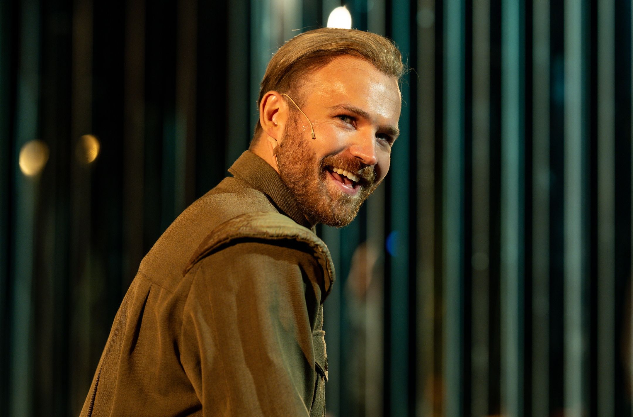 Smilende mann med mikrofon på kinnet ser mot kamera iført militærinspirert jakke.