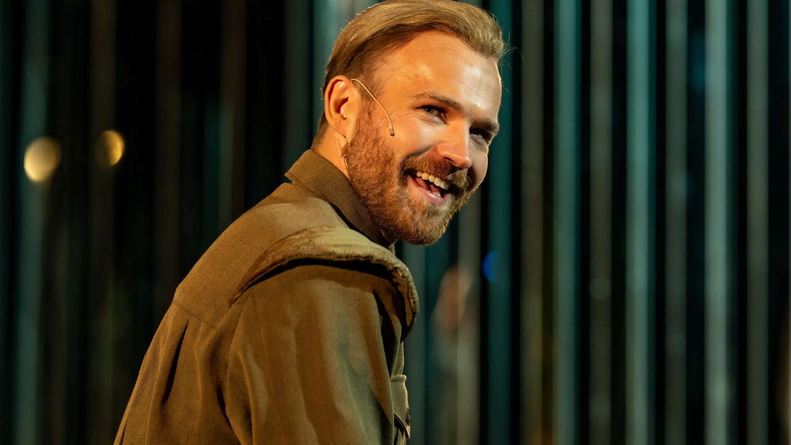 Smilende mann med mikrofon på kinnet ser mot kamera iført militærinspirert jakke.