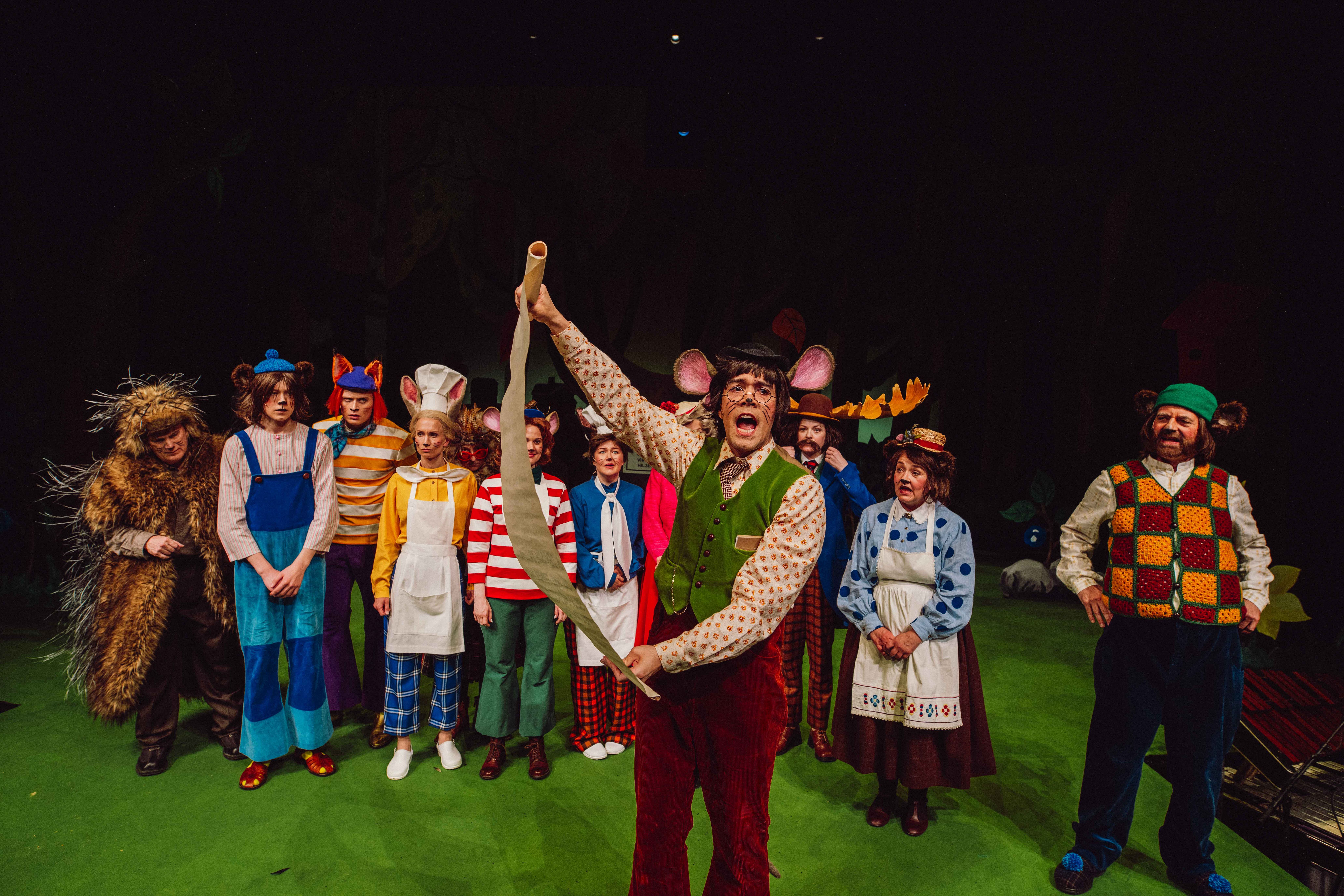 En mus med pergamentrull leser høyt foran en gruppe karakterer i dyrekostymer.