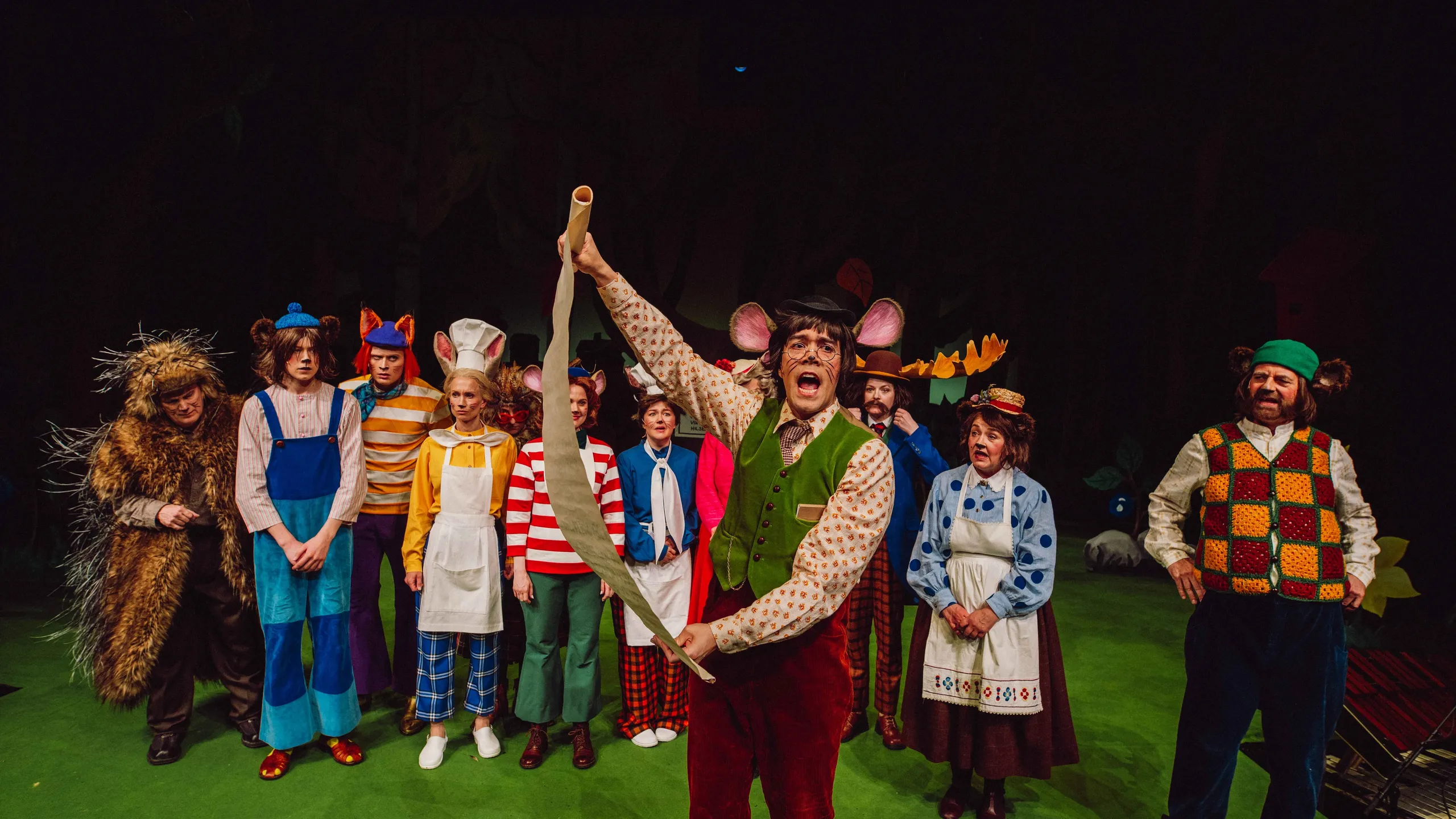 En mus med pergamentrull leser høyt foran en gruppe karakterer i dyrekostymer.