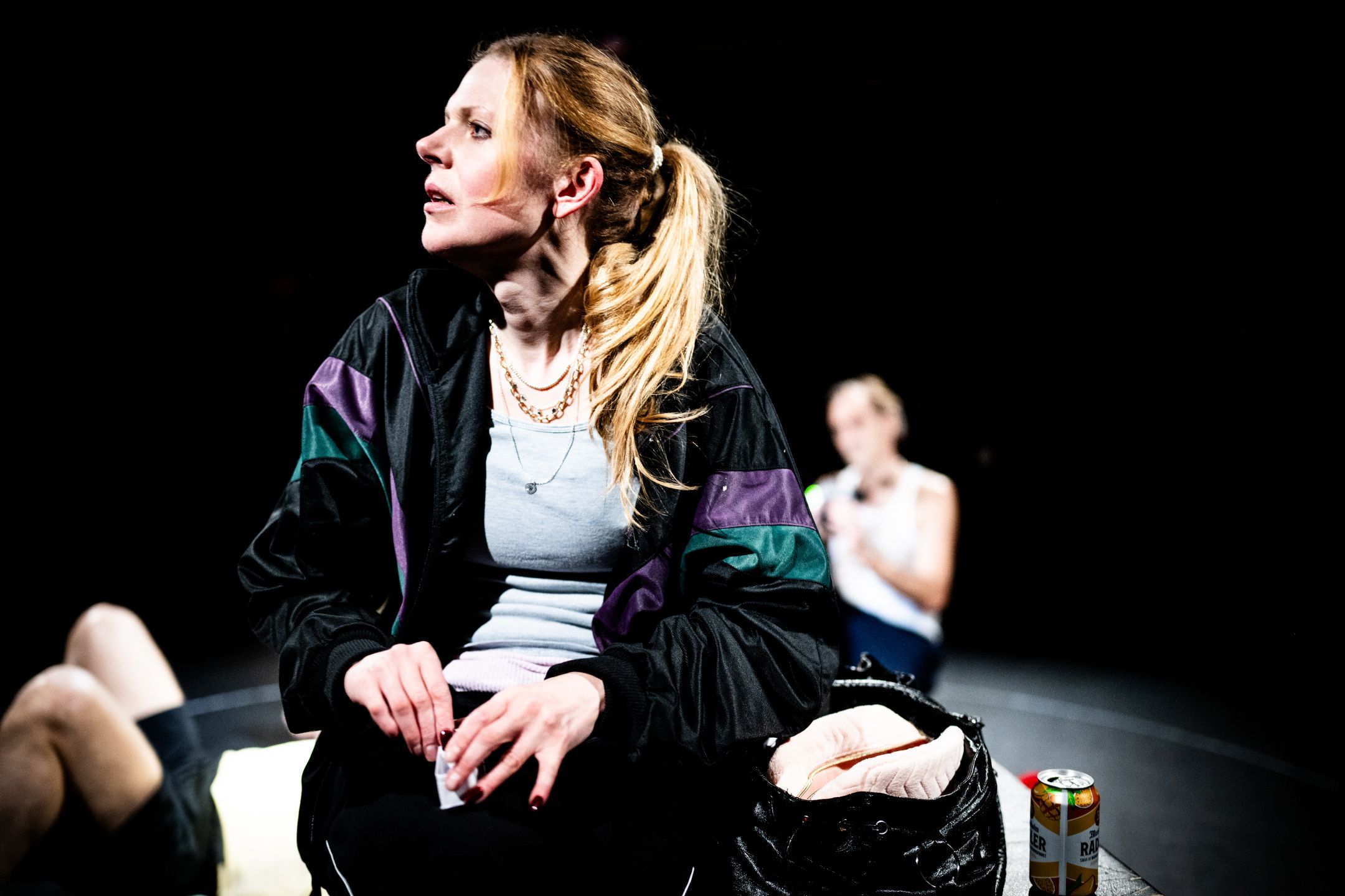 Kvinne i treningstøy sitter på scenekant og ser til siden, med ølboks og bag foran seg