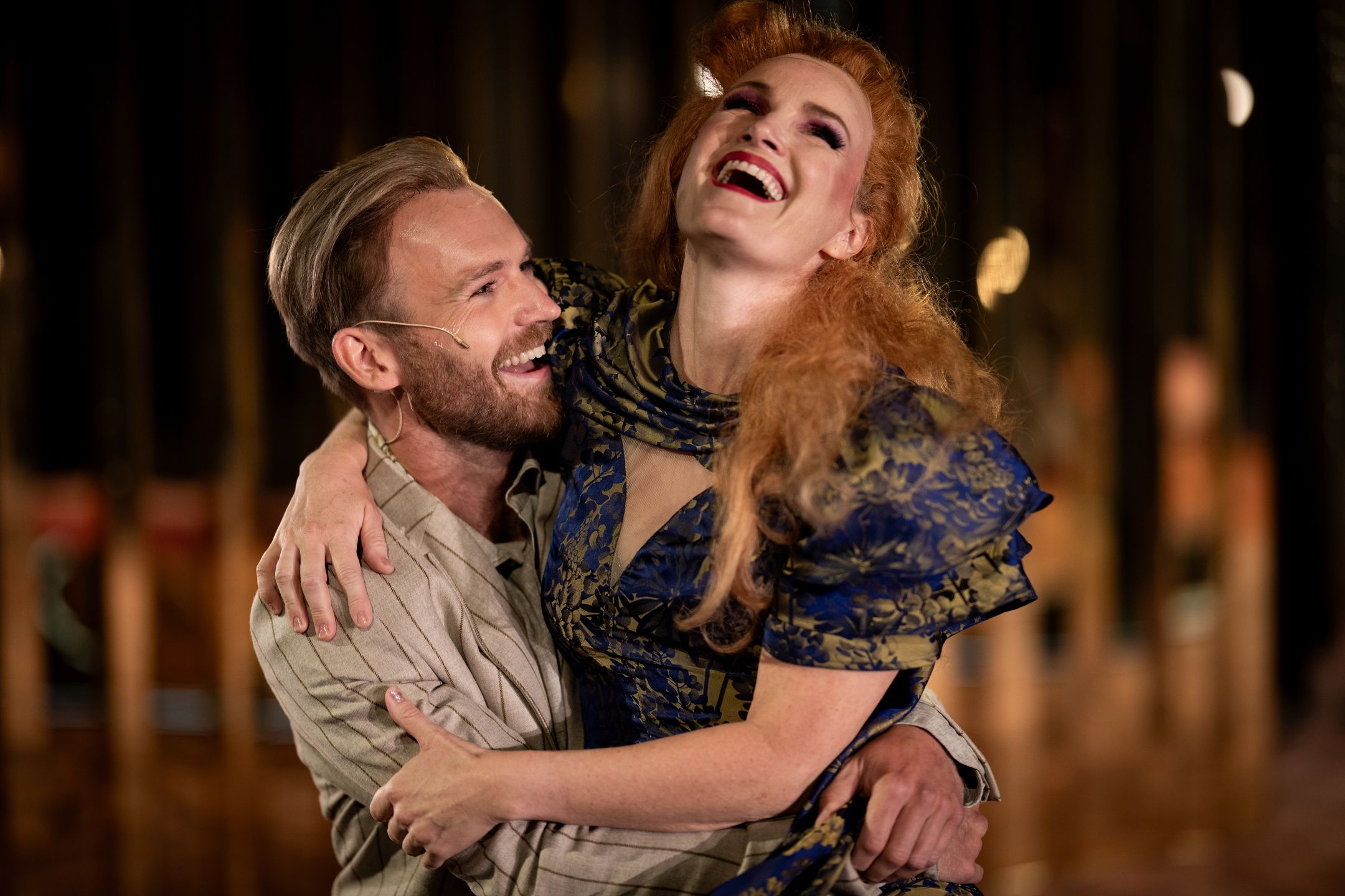 En mann og kvinne i teaterkostymer ler sammen, kvinnen i blå og gull kjole løftes i mannens armer.