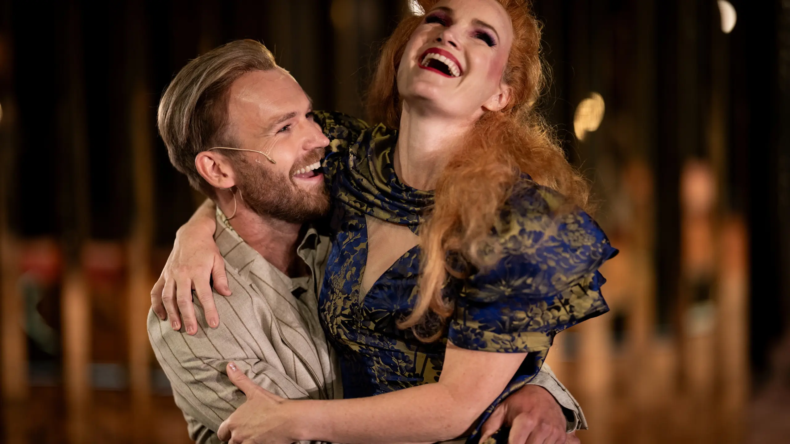 En mann og kvinne i teaterkostymer ler sammen, kvinnen i blå og gull kjole løftes i mannens armer.