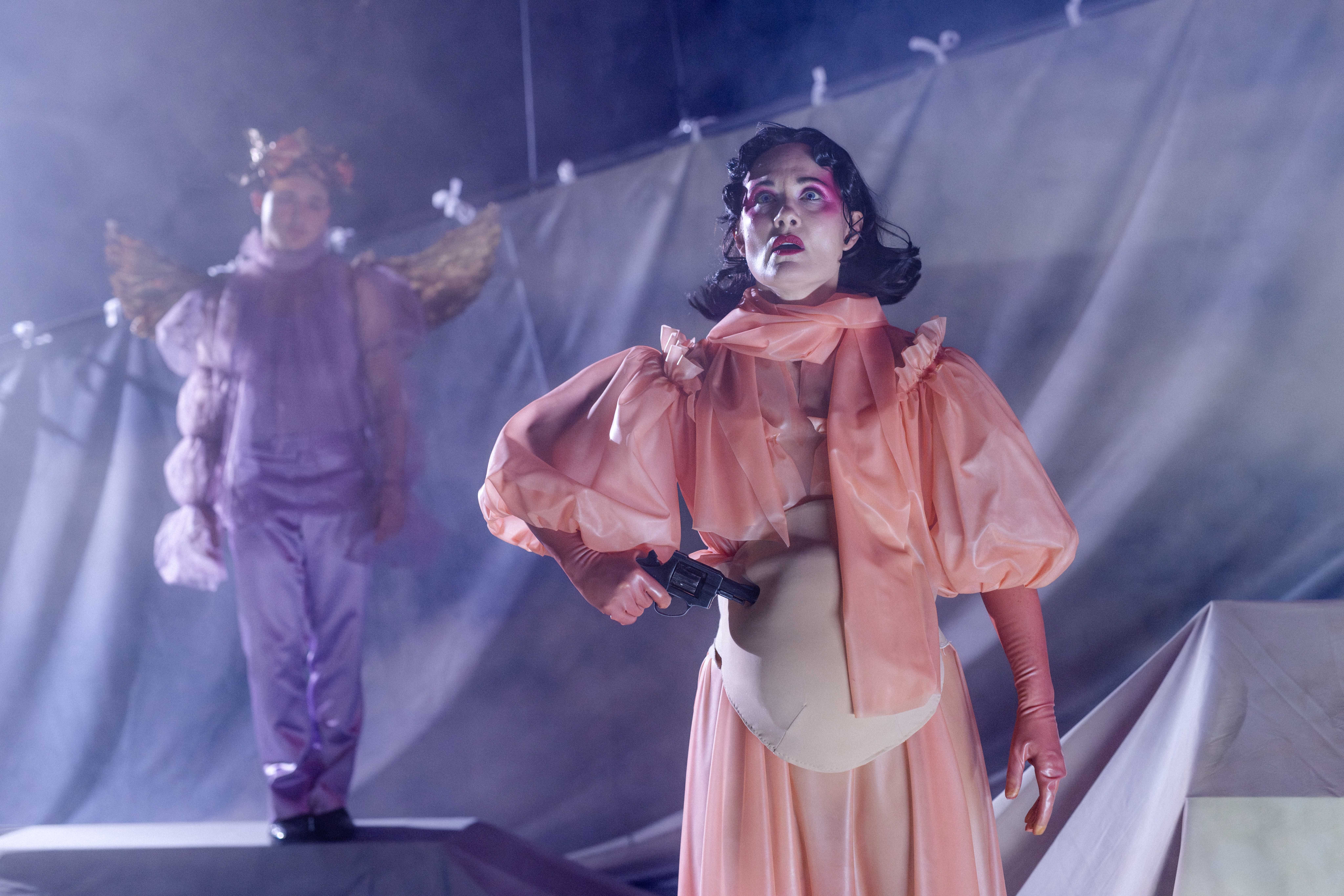 Person i ferskenfarget kjole står på scenen og holder en pistol mot magen, mens en figur med vinger står uklart i bakgrunnen