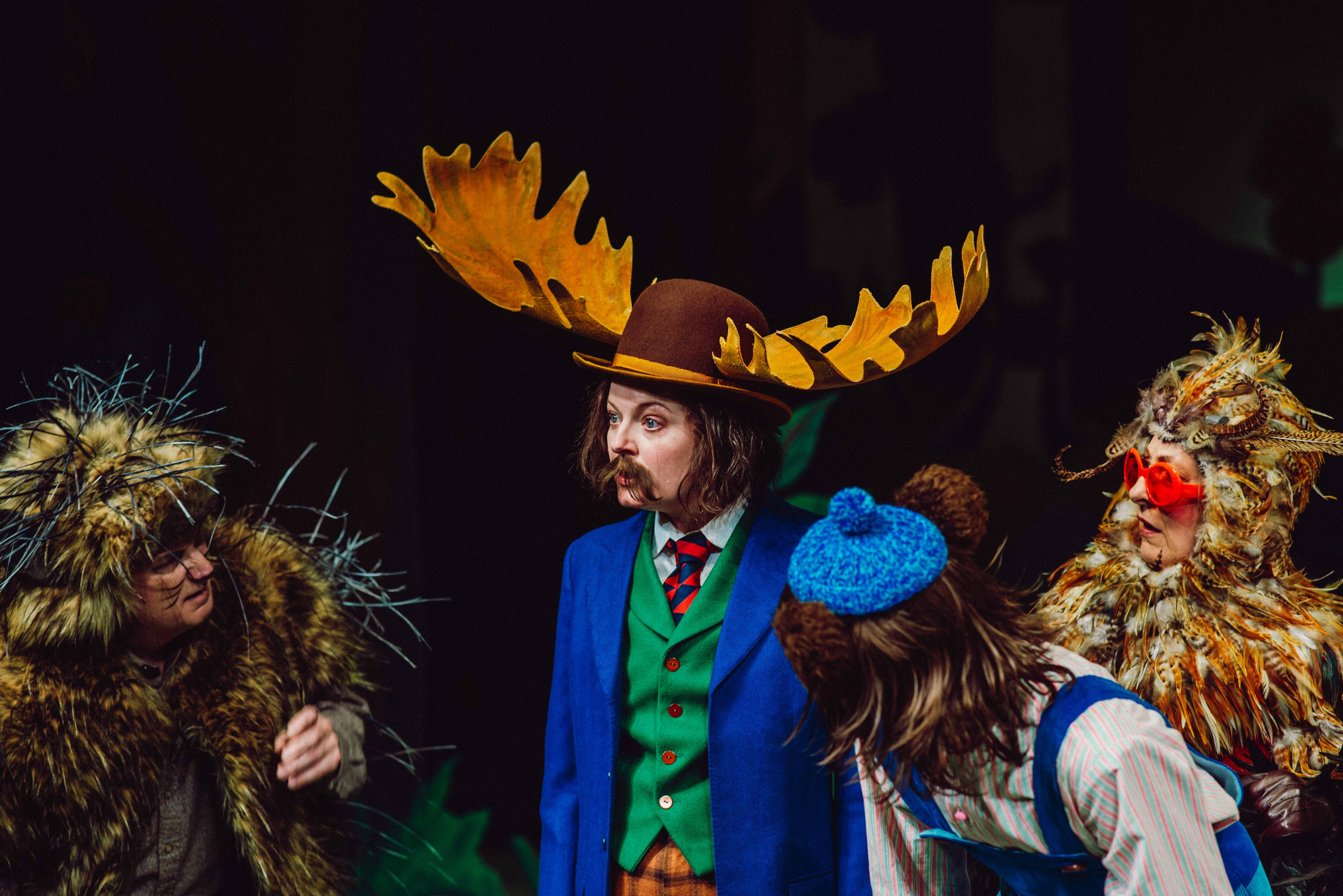 Fire personer i dyrekostymer står samlet, med én i midten iført blå jakke og stor brun hatt med elggevir.