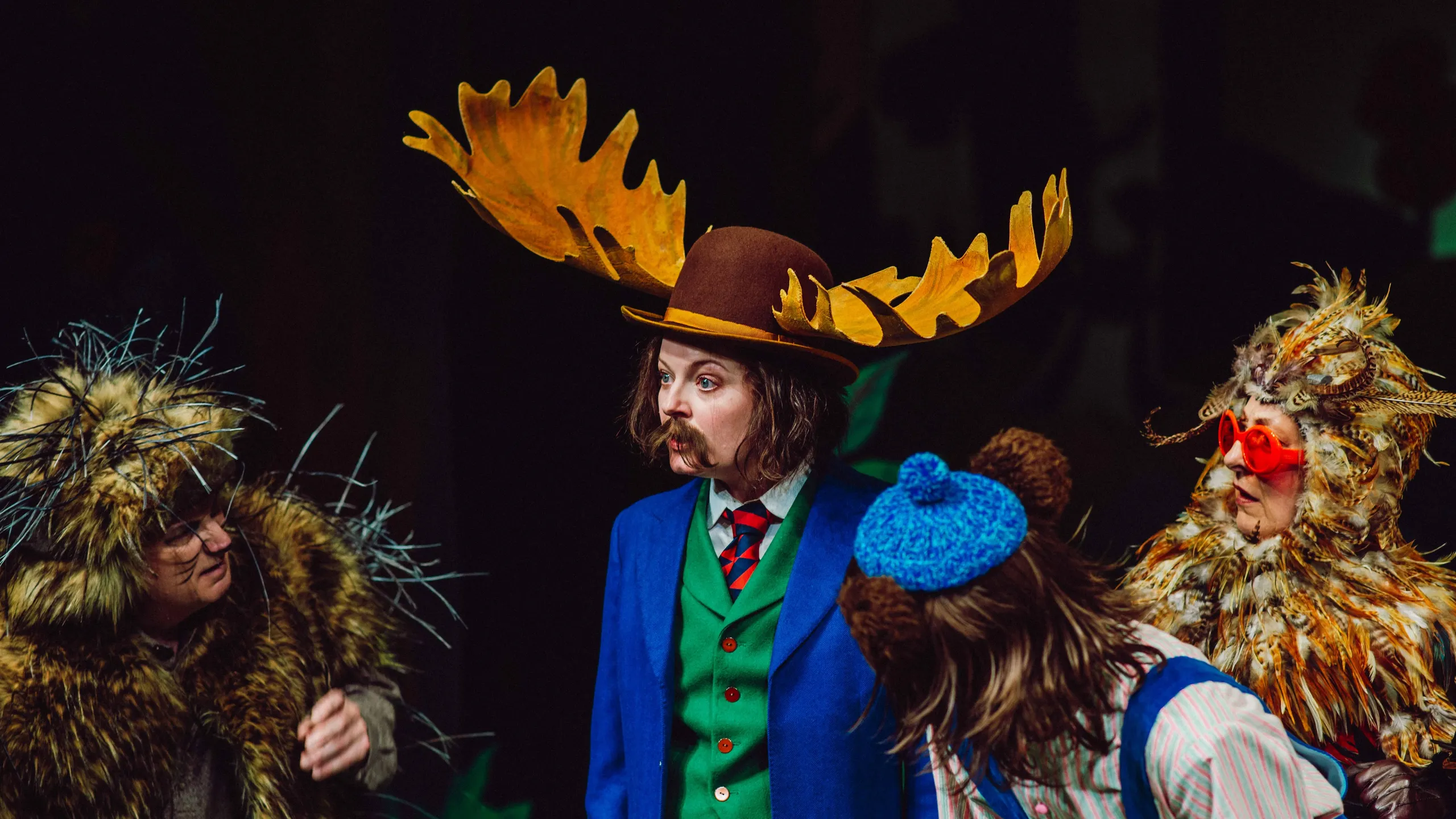 Fire personer i dyrekostymer står samlet, med én i midten iført blå jakke og stor brun hatt med elggevir.