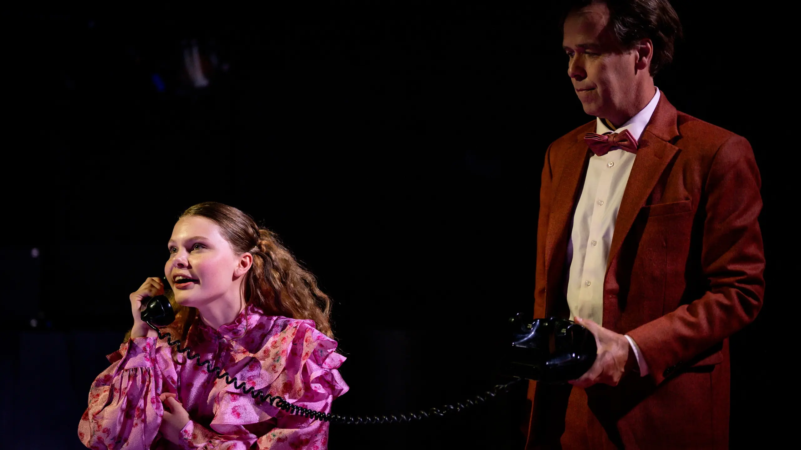 Jente i rosa blomstret bluse snakker i telefon mens en mann i rød dress og sløyfe holder apparatet, begge står i mørkt scenerom.