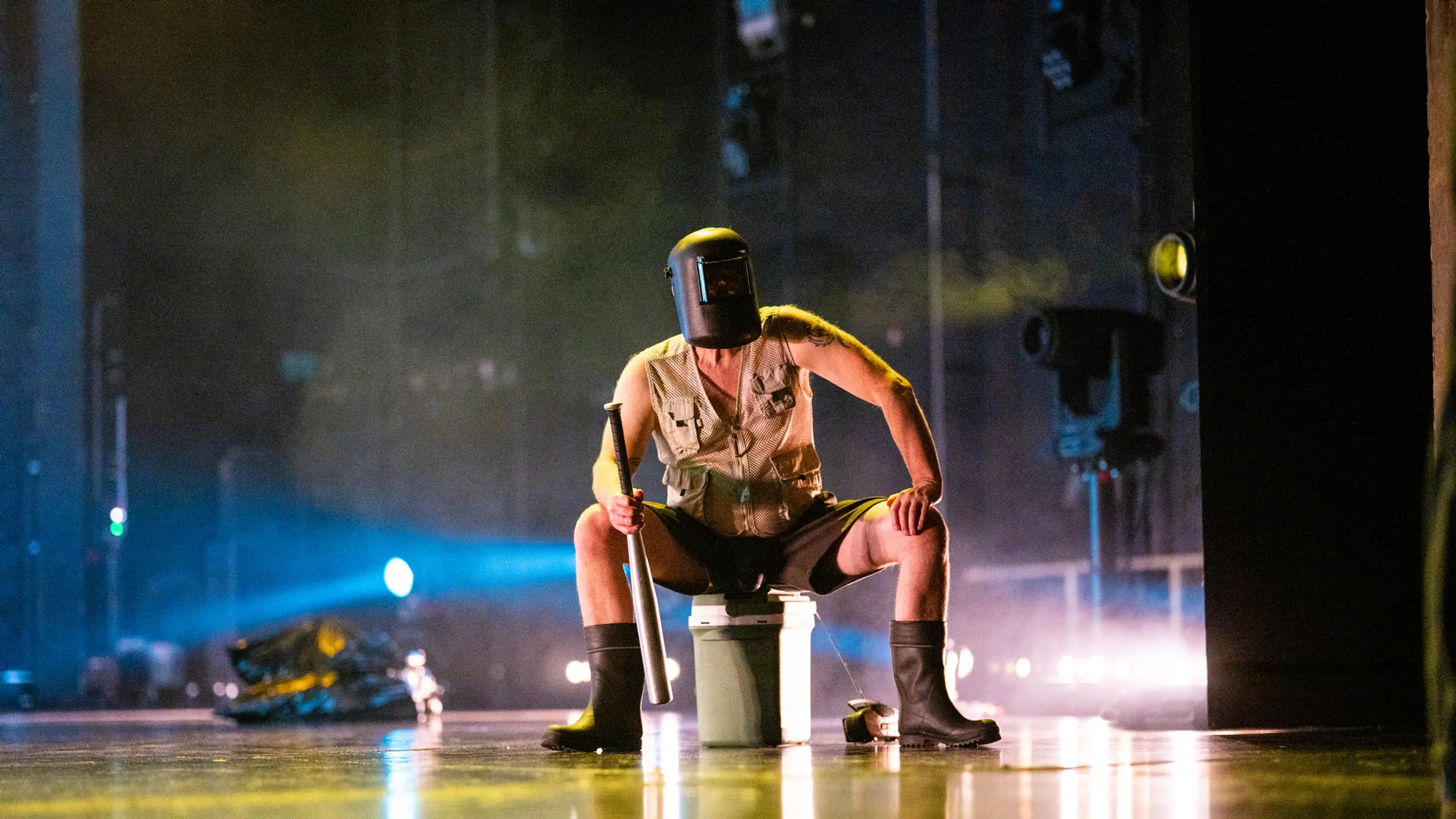Skuespiller i sveisehjelm og vest sitter bredbeint på bøtte med fiskestang, iført støvler og shorts.