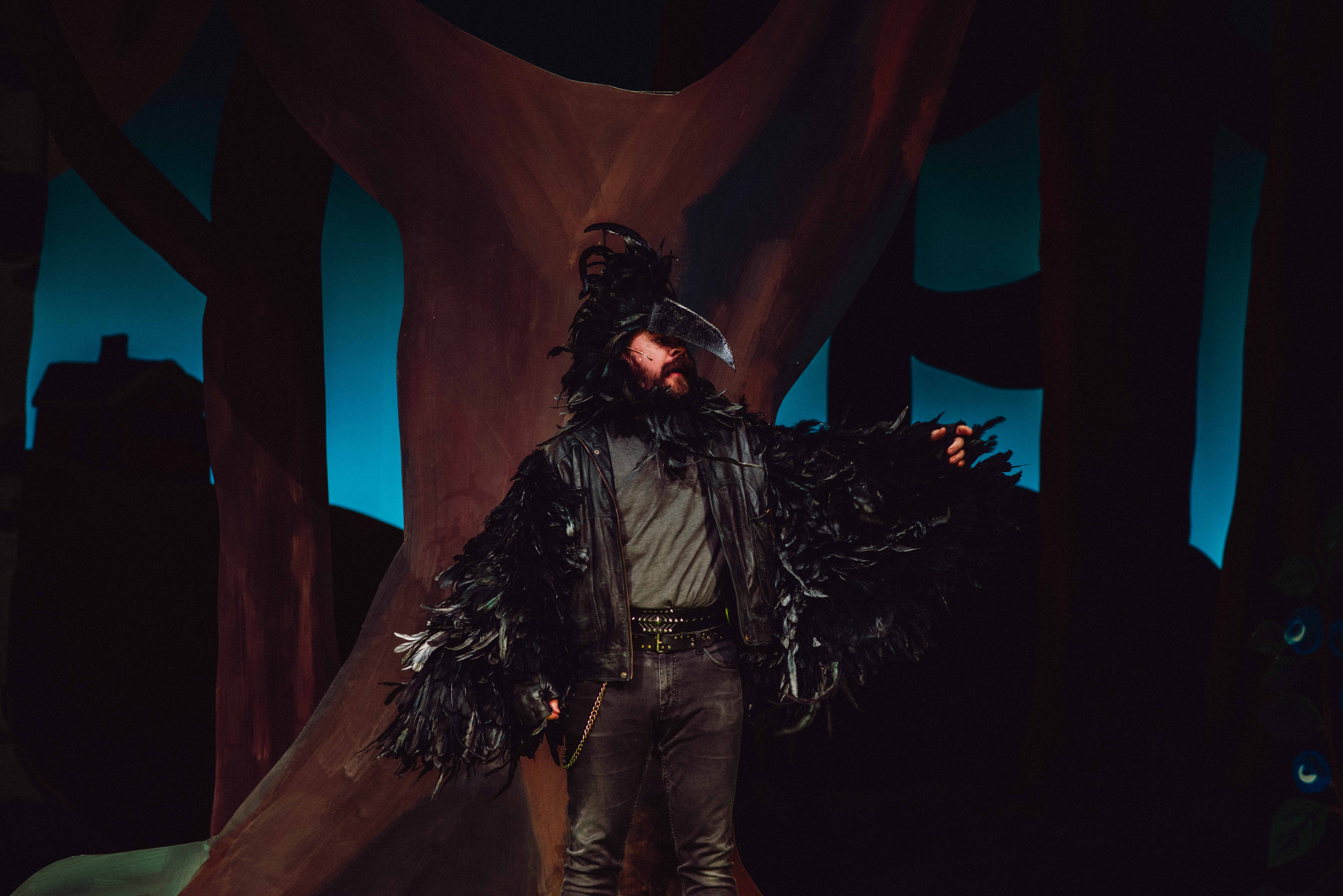 Person i svart fjærdrakt og nebb står foran trestamme med mørk bakgrunn.