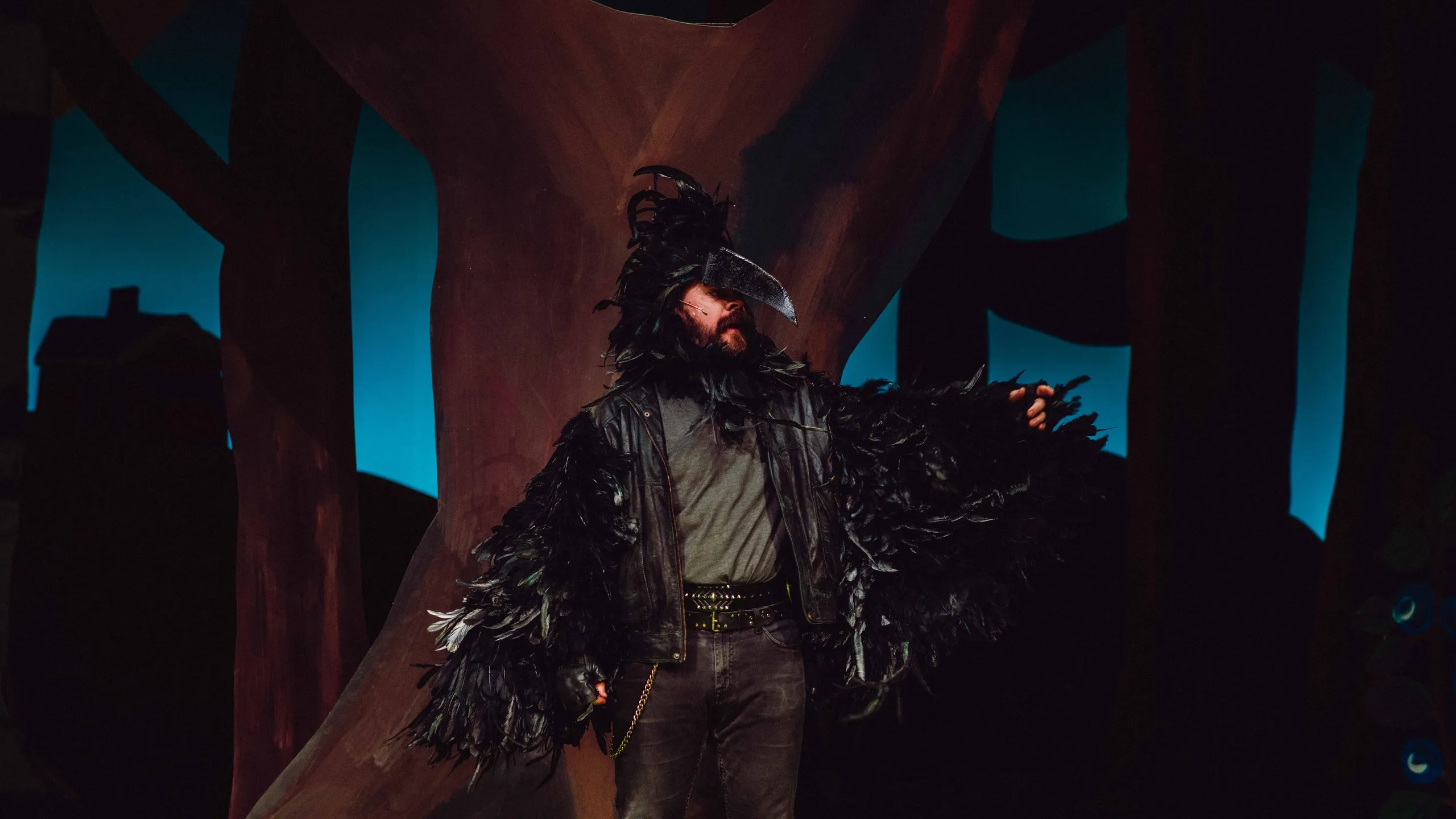 Person i svart fjærdrakt og nebb står foran trestamme med mørk bakgrunn.