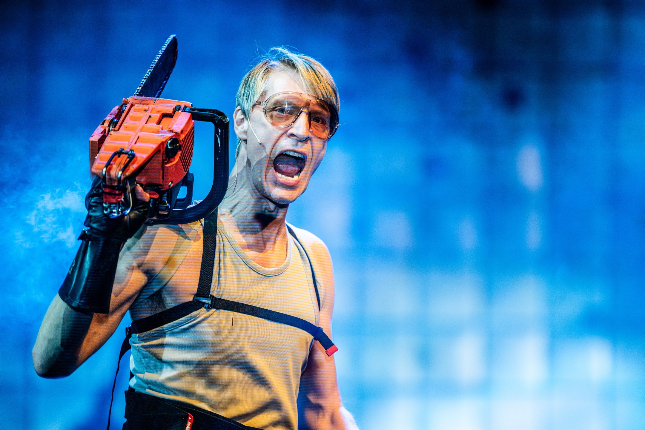 Person med lys hud og briller roper mens han holder en motorsag mot blå bakgrunn.