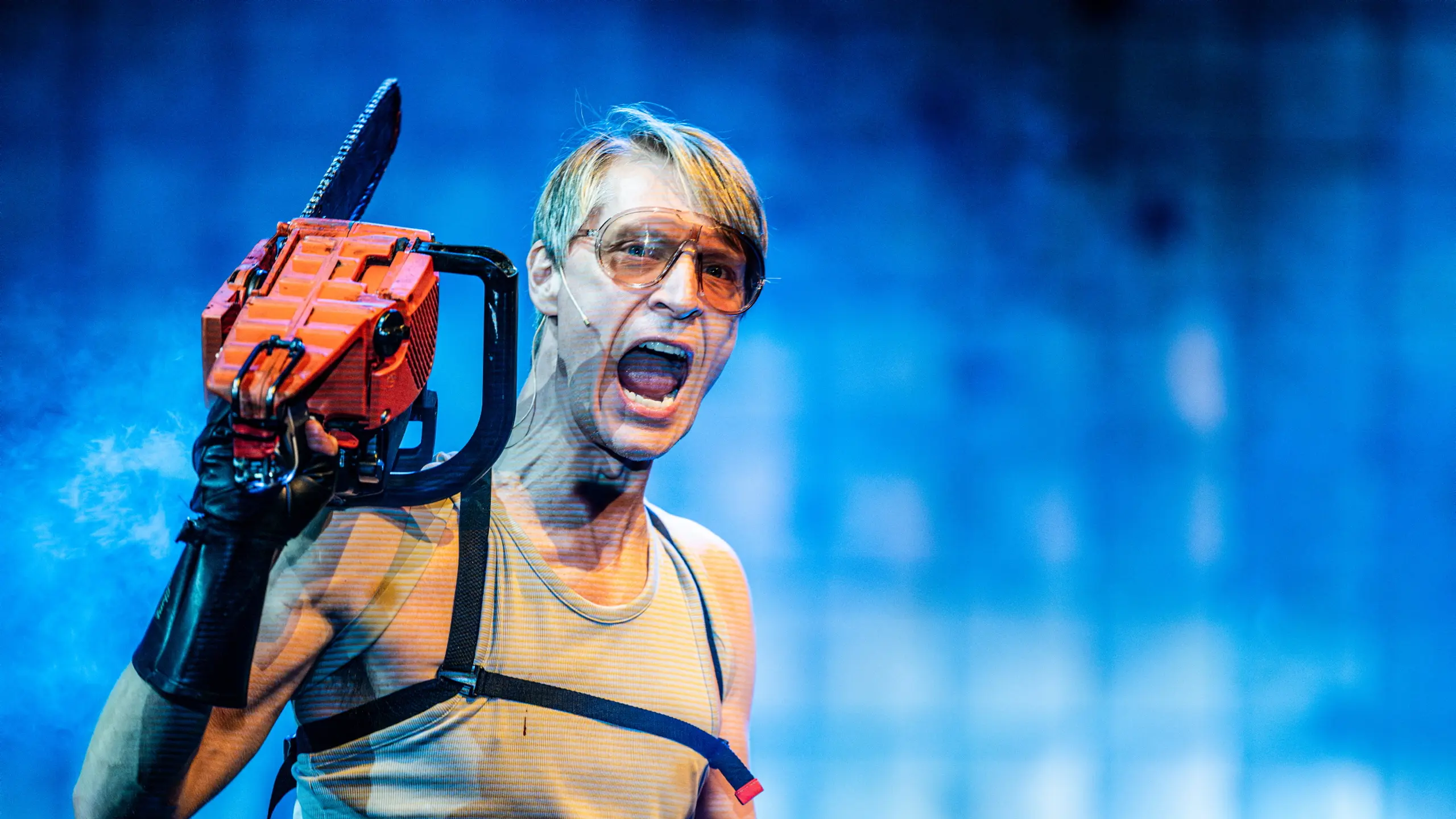 Person med lys hud og briller roper mens han holder en motorsag mot blå bakgrunn.