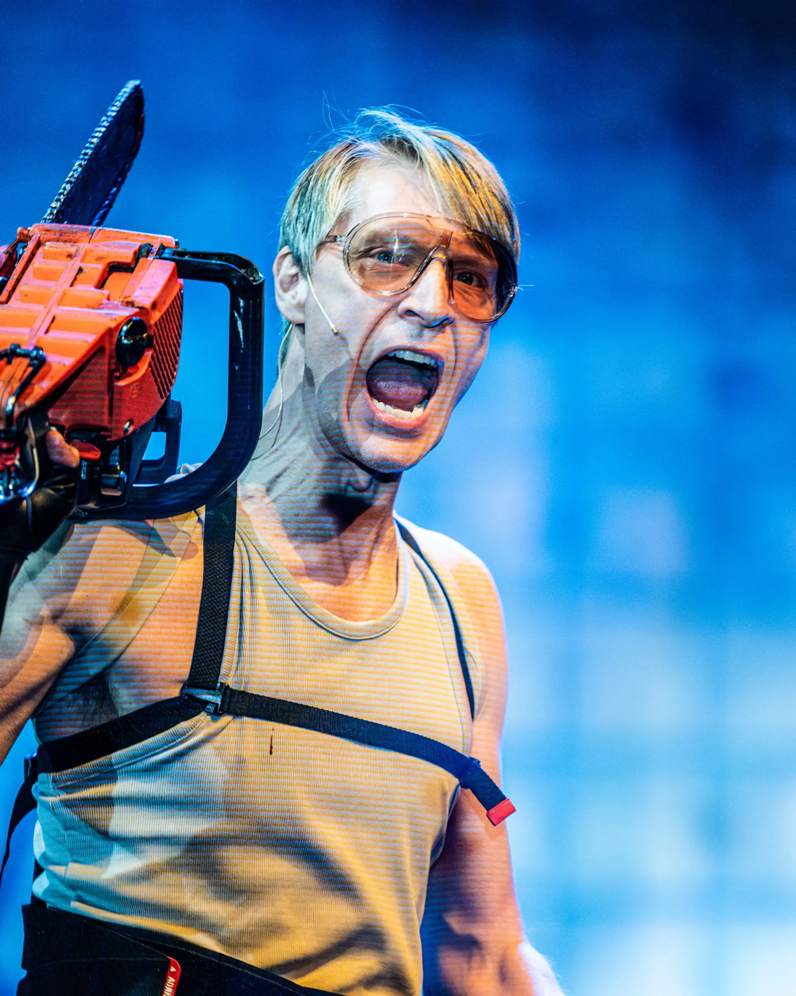 Person med lys hud og briller roper mens han holder en motorsag mot blå bakgrunn.