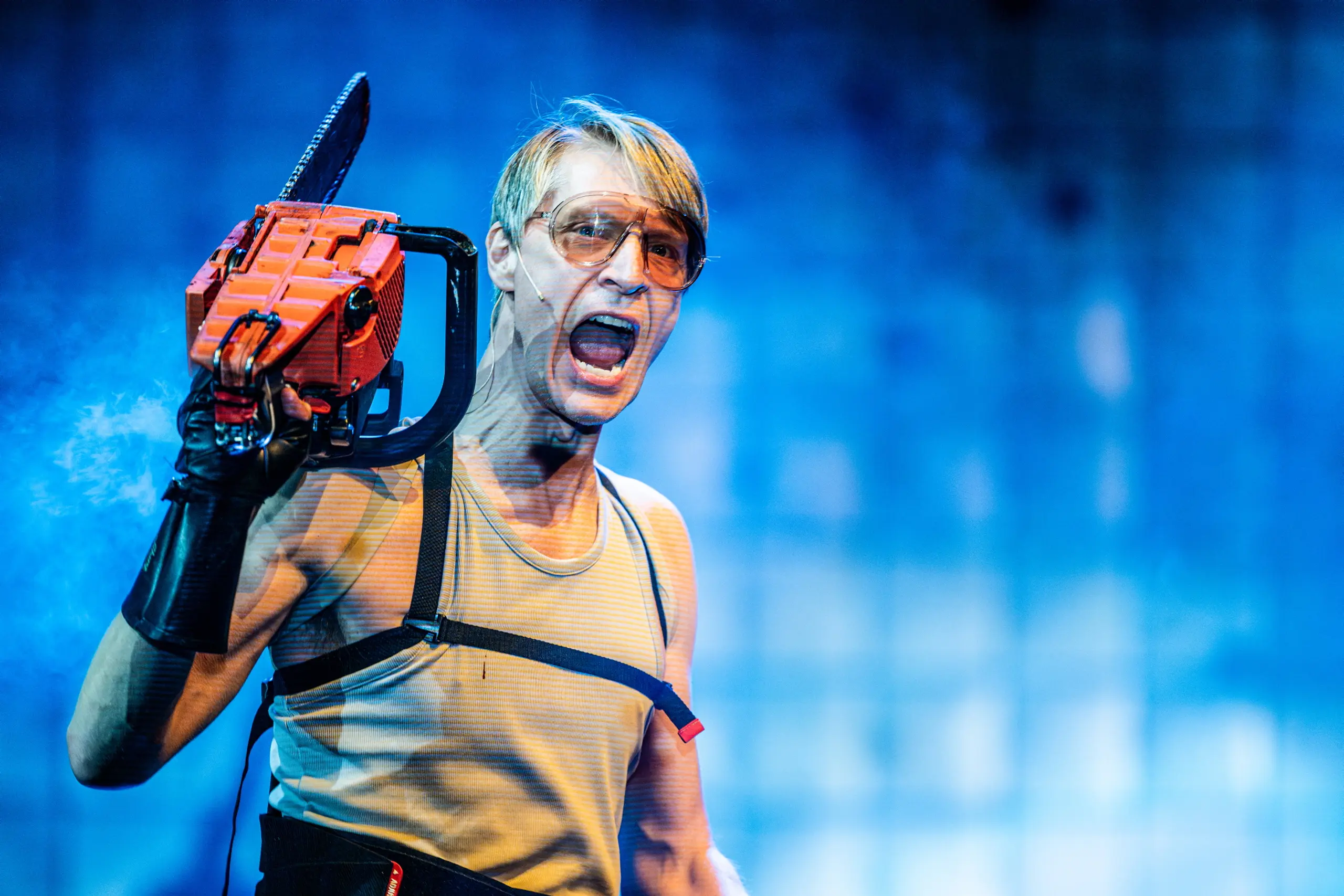 Person med lys hud og briller roper mens han holder en motorsag mot blå bakgrunn.