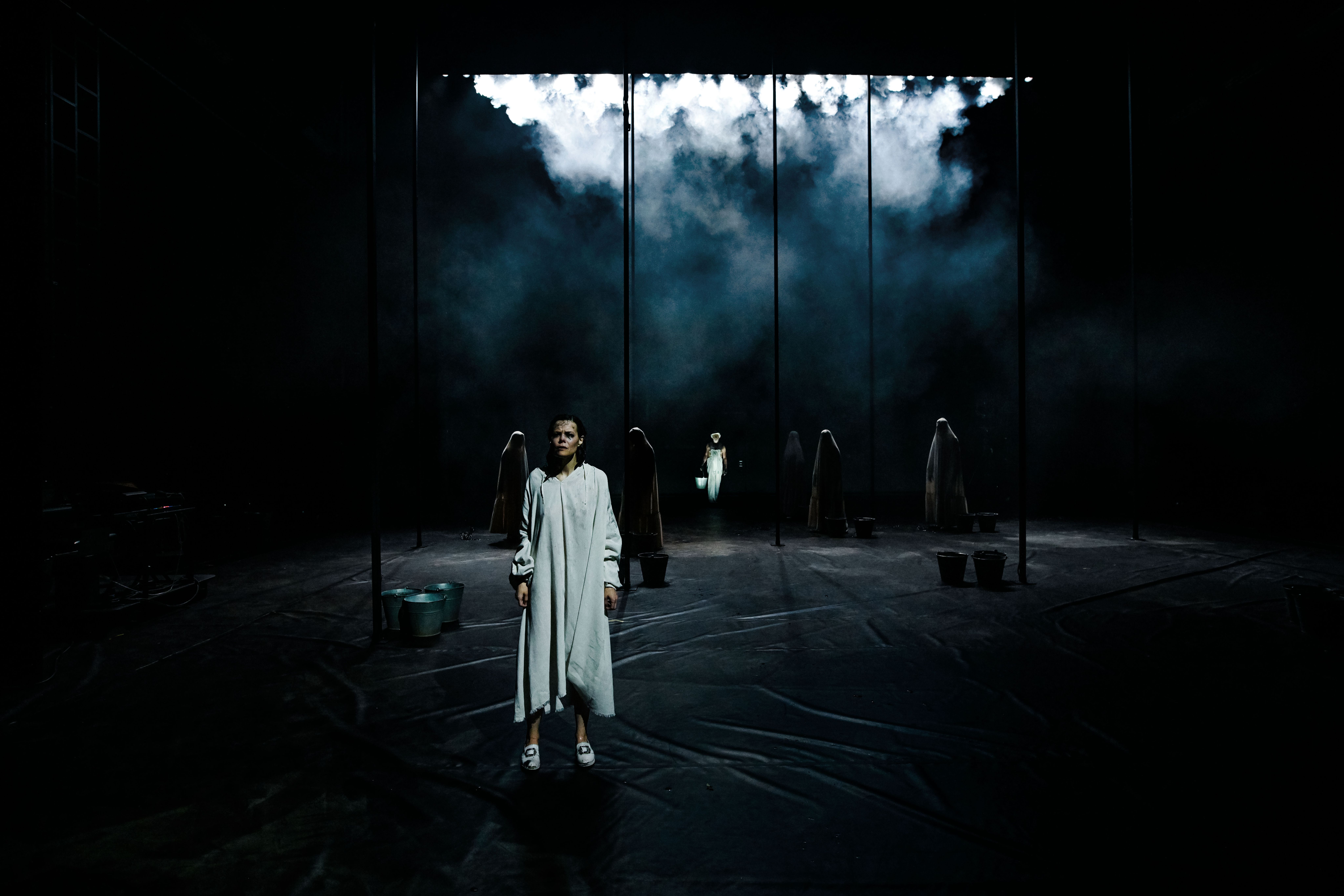 En kvinne står i forgrunnen på en mørk, røykfylt scene. Hun har hvit kjortel og hvite bunadssko. I bakgrunnen ser vi hvite skikkelser med tildekket hode og bøtter