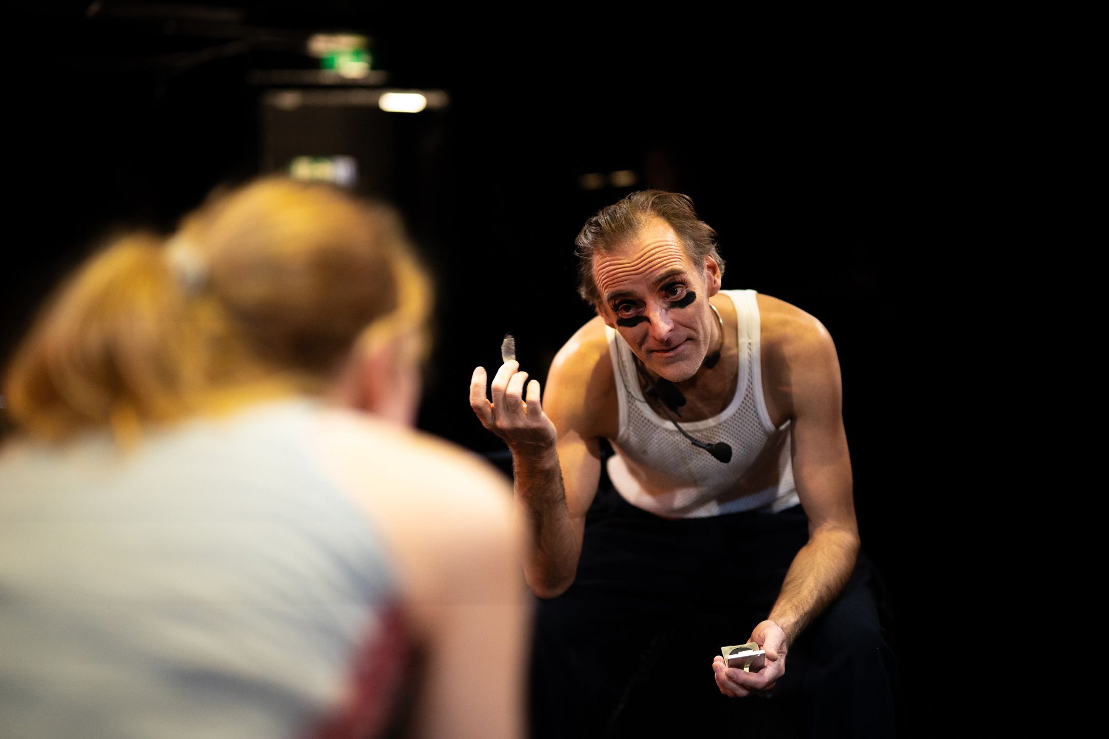 Mann i hvit nettingtrøye med svart ansiktsmaling holder noe lite i hånden og ser mot en kvinne i forgrunnen