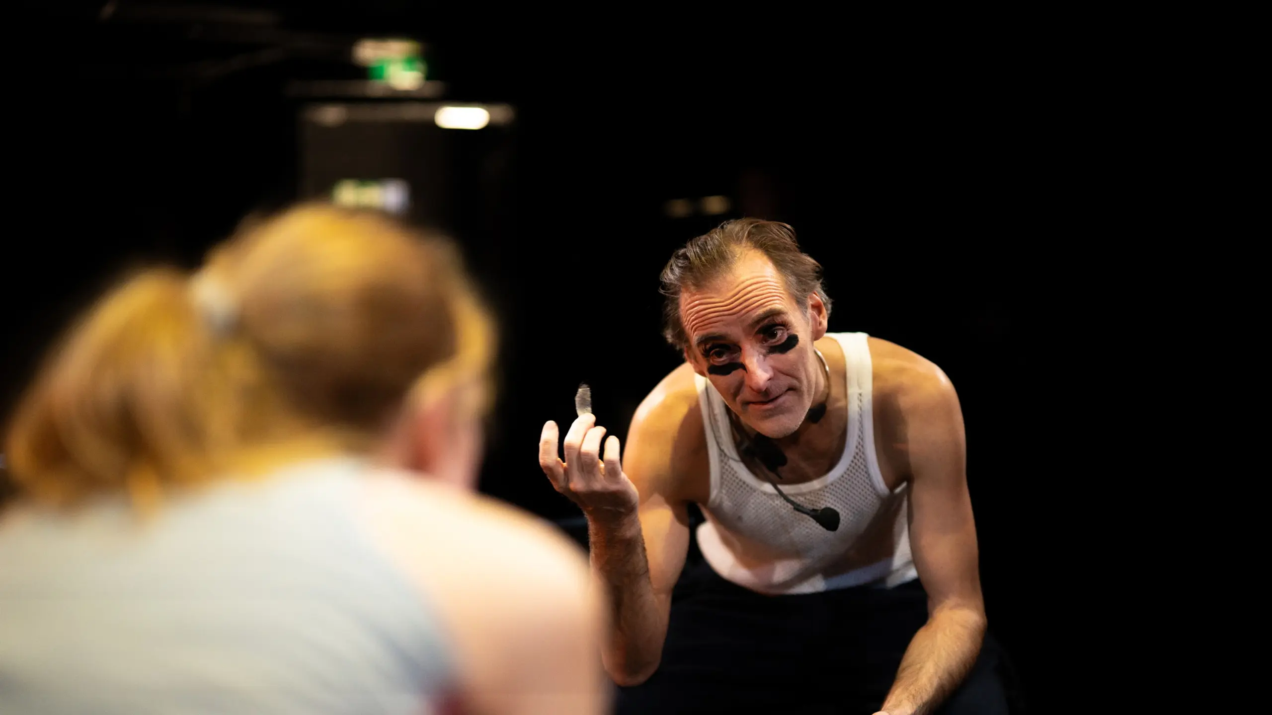 Mann i hvit nettingtrøye med svart ansiktsmaling holder noe lite i hånden og ser mot en kvinne i forgrunnen