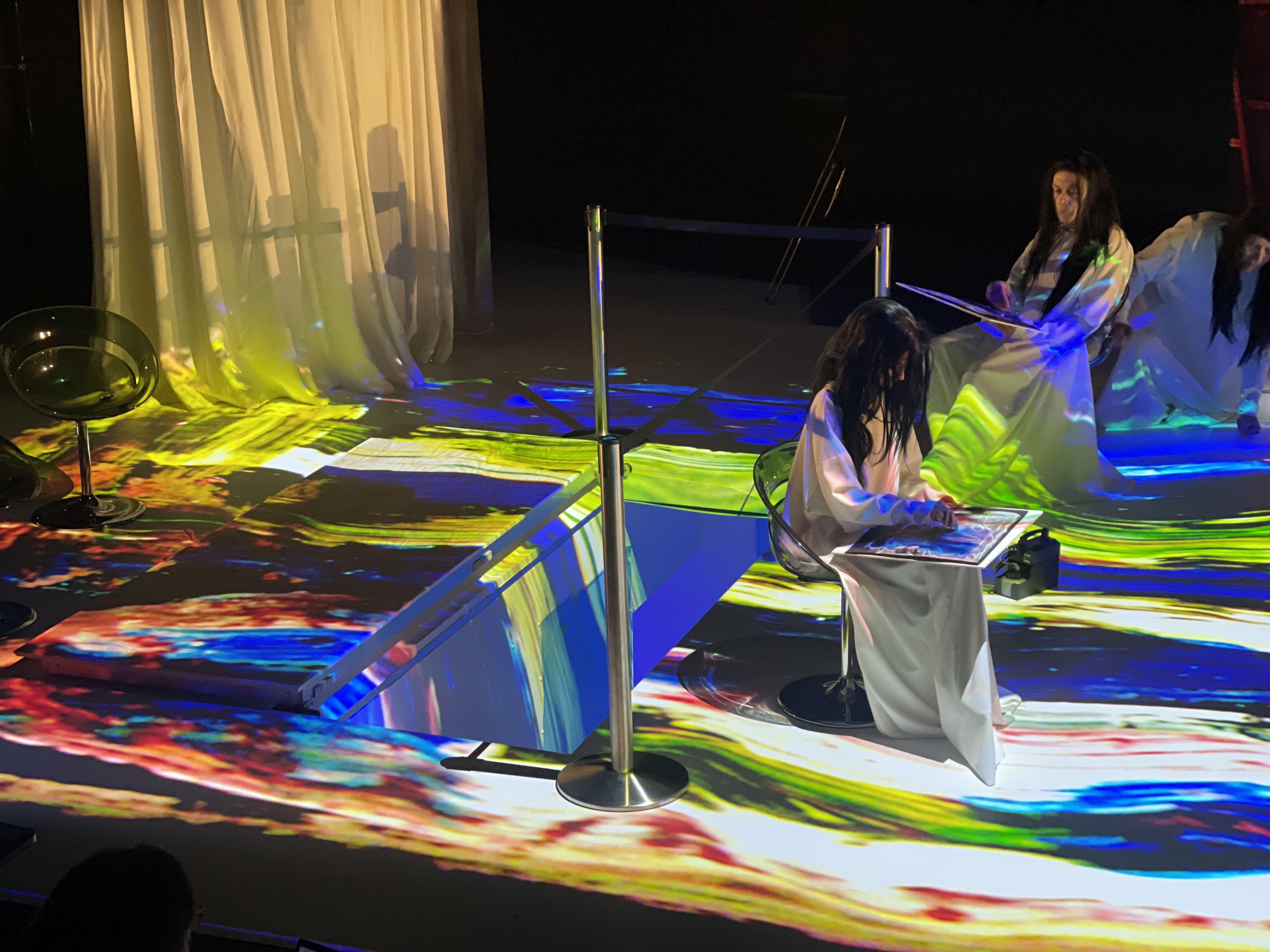 Prøvebilder fra scenen på forestillingen Kallocain - vi ser to skuespillere i hvite skjortler og med parykker mad langt sort hår. De er badet i fargerikt lys og holder i noe som ser ut som store, flate skjermer