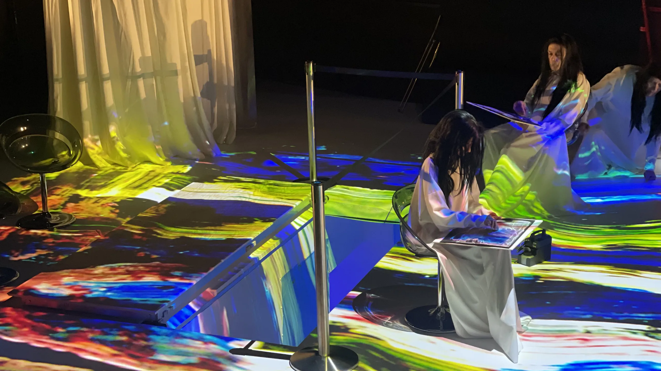 Prøvebilder fra scenen på forestillingen Kallocain - vi ser to skuespillere i hvite skjortler og med parykker mad langt sort hår. De er badet i fargerikt lys og holder i noe som ser ut som store, flate skjermer