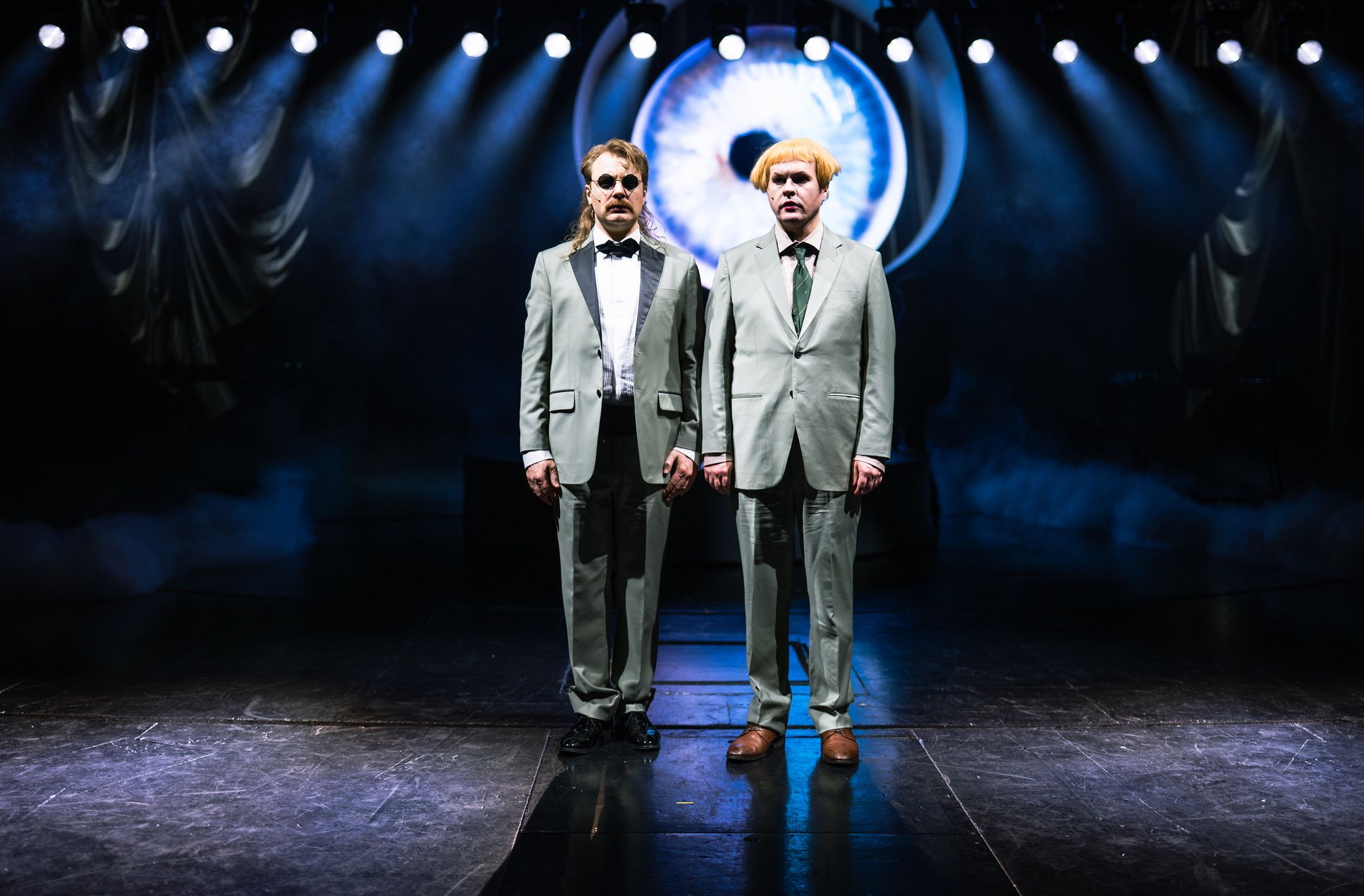 To personer i matchende lysegrå dresser står rett opp og ned på scenen under spottlys foran et digitalt øyemotiv.