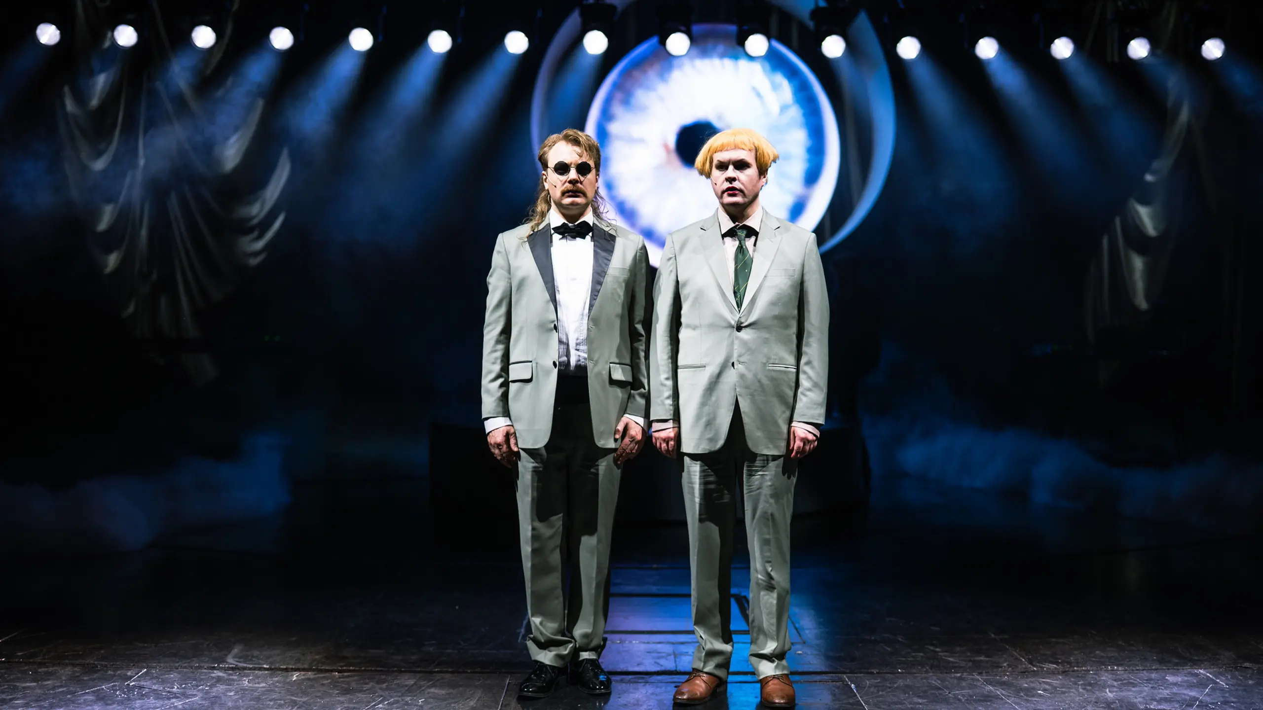 To personer i matchende lysegrå dresser står rett opp og ned på scenen under spottlys foran et digitalt øyemotiv.