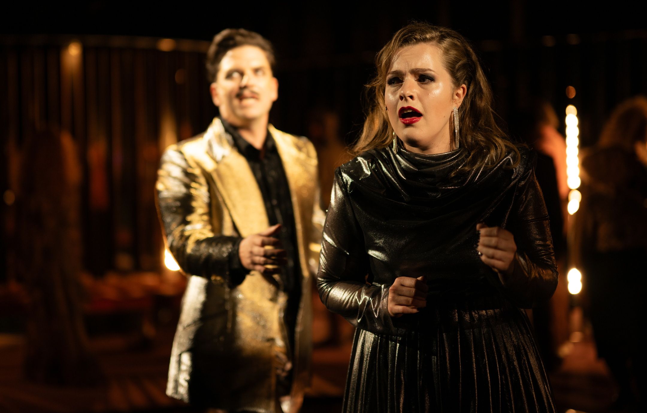 Kvinne i skinnende mørk kjole synger, mens mann i gulljakke står i bakgrunnen i uklart fokus.