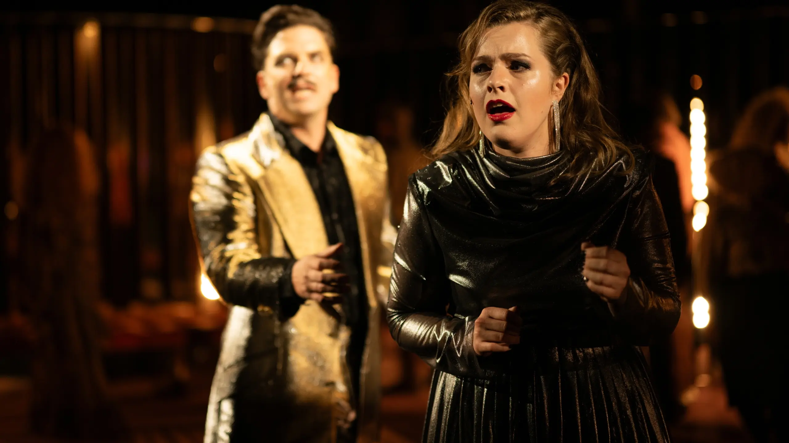 Kvinne i skinnende mørk kjole synger, mens mann i gulljakke står i bakgrunnen i uklart fokus.
