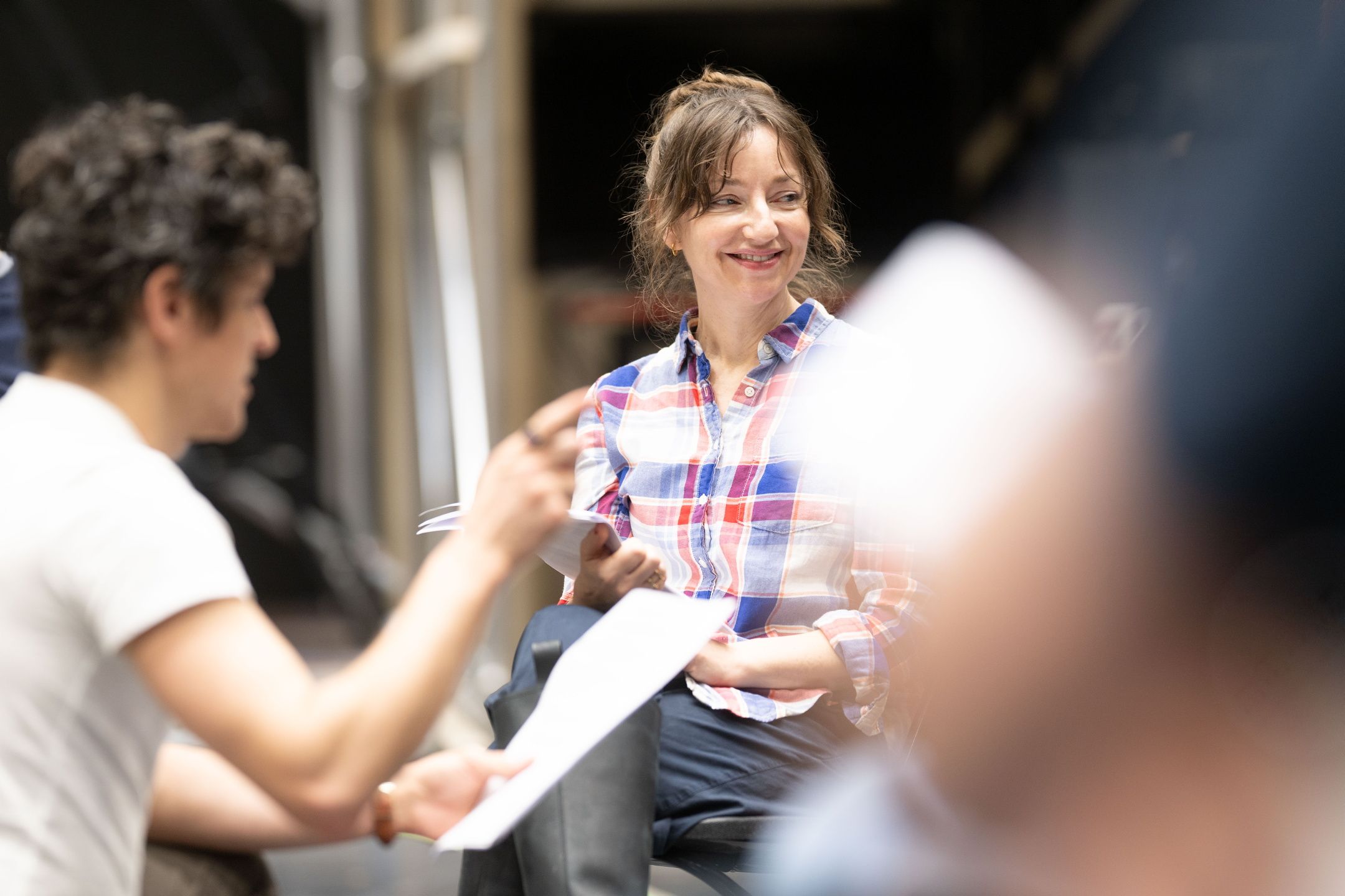 En kvinne i rutete skjorte smiler under en bordprøve mens hun holder manus, omgitt av andre skuespillere.