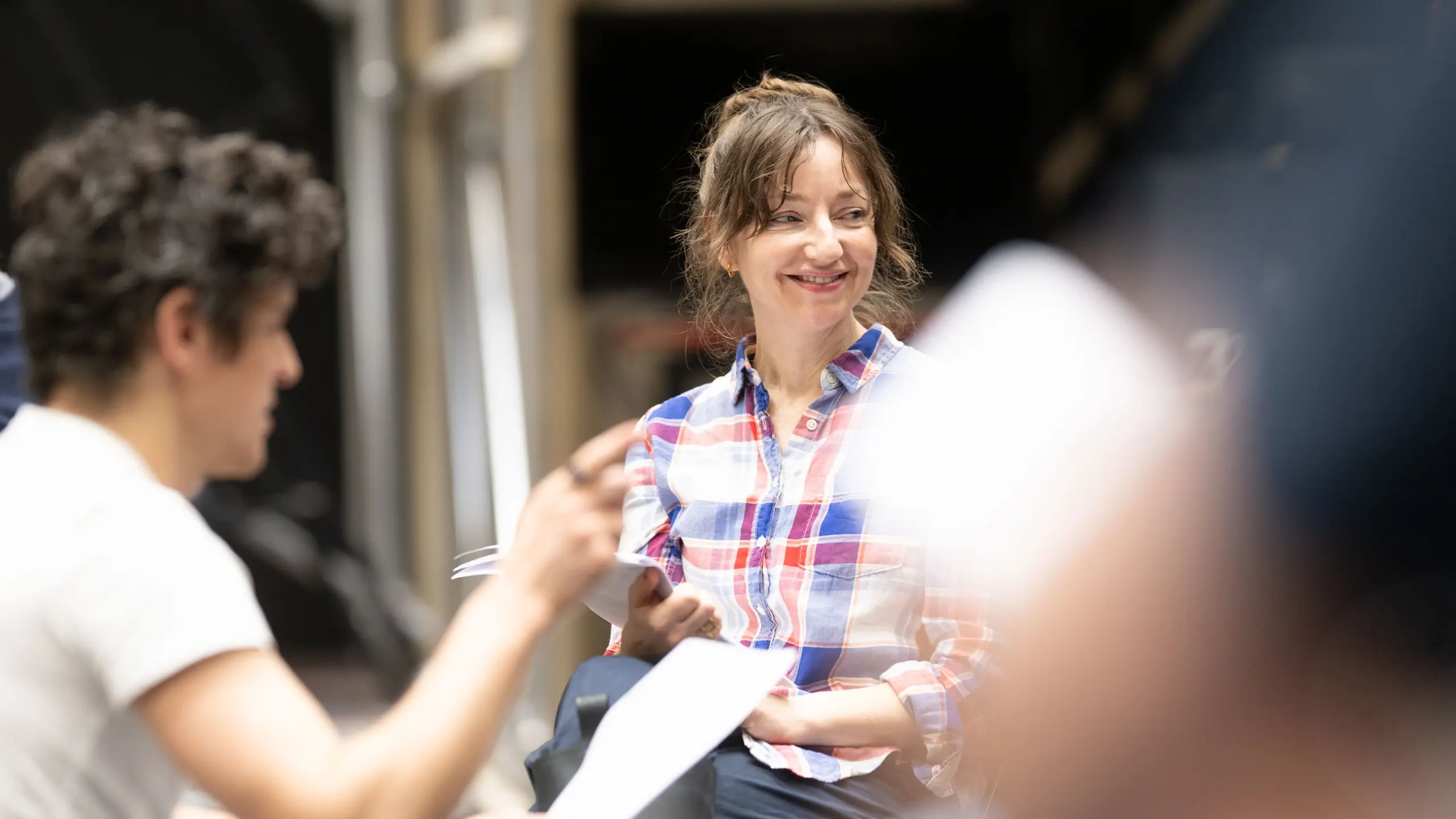 En kvinne i rutete skjorte smiler under en bordprøve mens hun holder manus, omgitt av andre skuespillere.