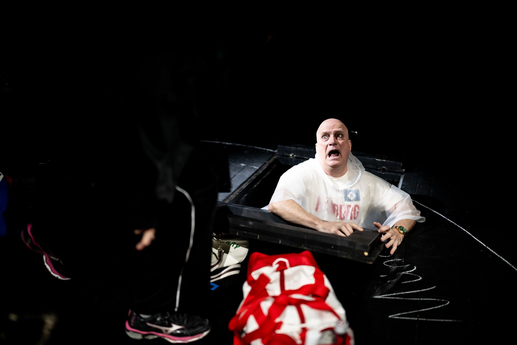 Mann i hvit poncho ser opp fra en luke i scenegulvet med forskrekket uttrykk