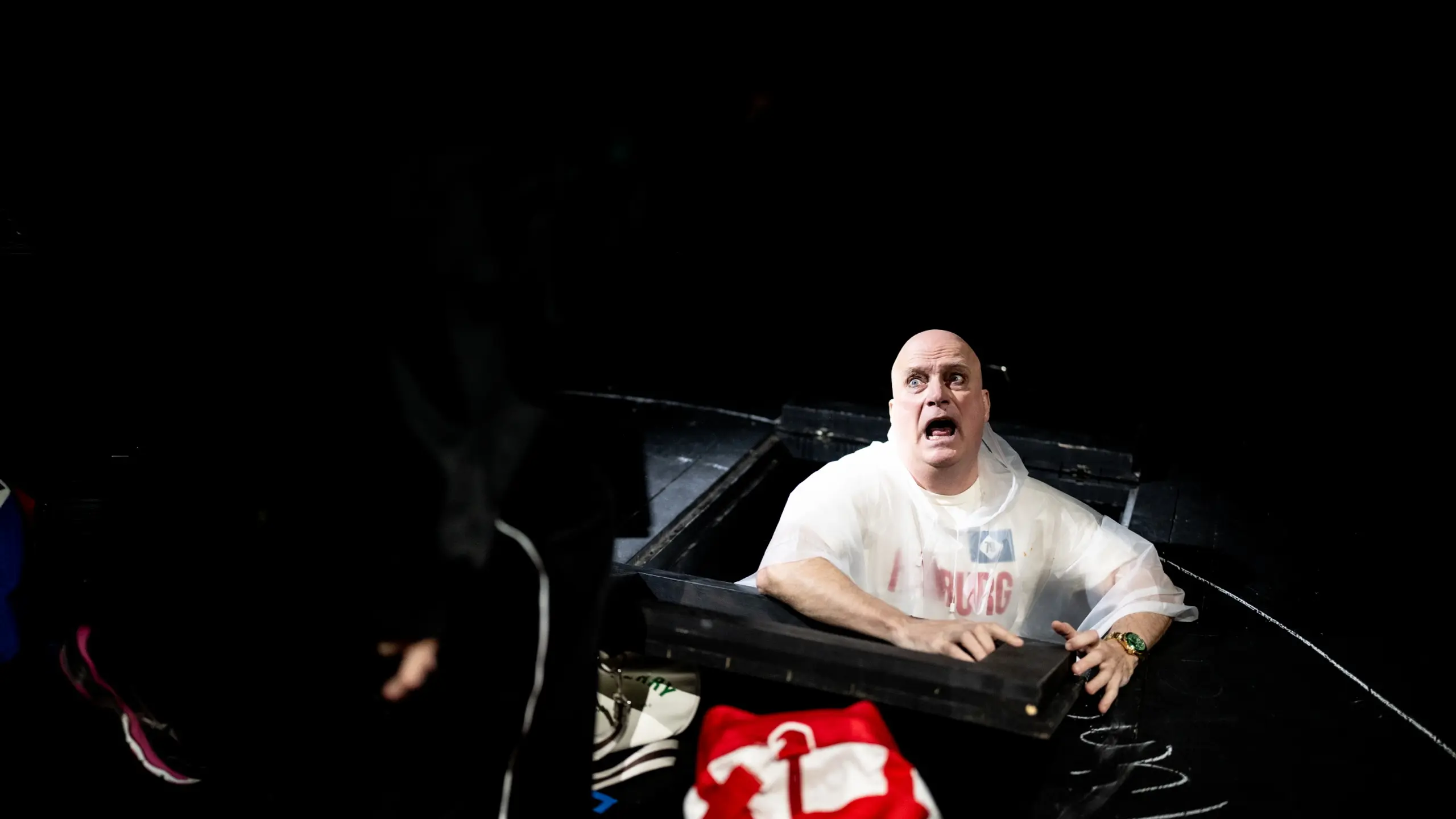 Mann i hvit poncho ser opp fra en luke i scenegulvet med forskrekket uttrykk