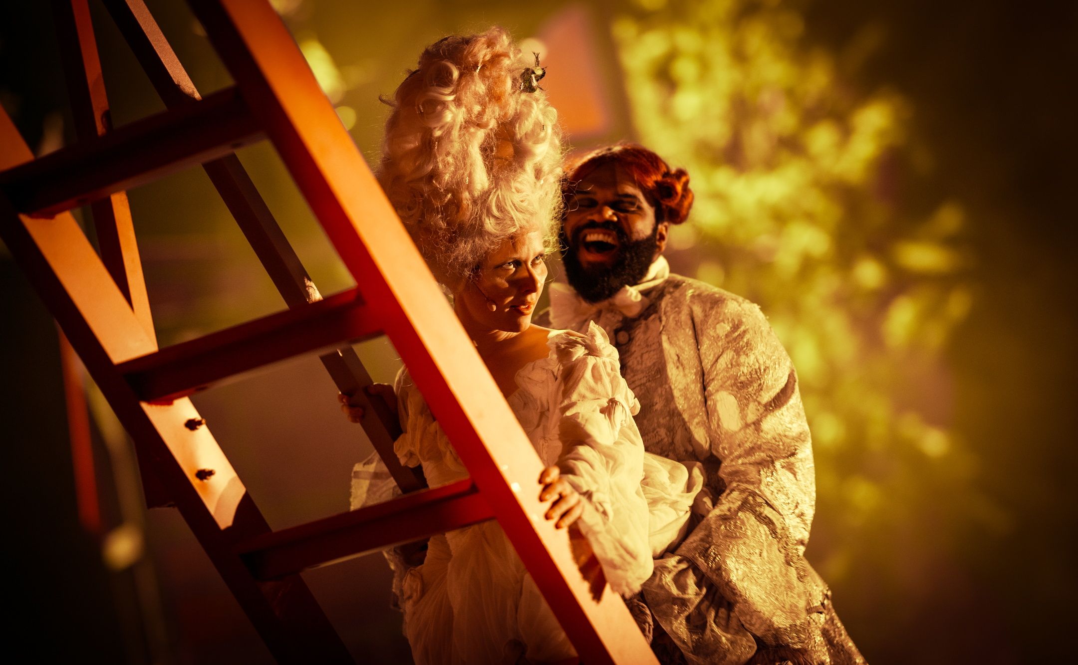 To skuespillere i overdådige, historiske kostymer smiler mens de står ved en rød stige i varmt scenelys.