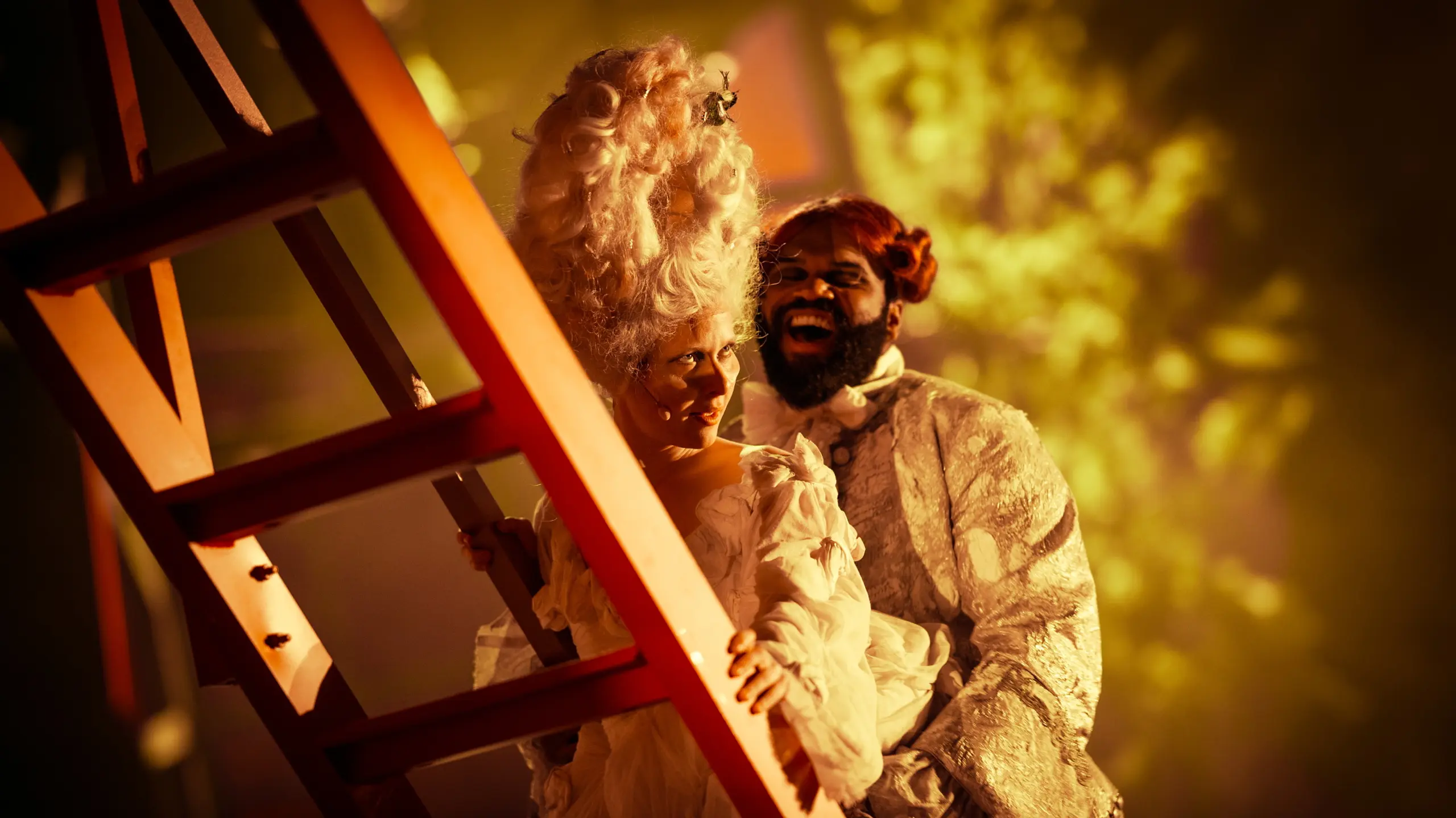 To skuespillere i overdådige, historiske kostymer smiler mens de står ved en rød stige i varmt scenelys.