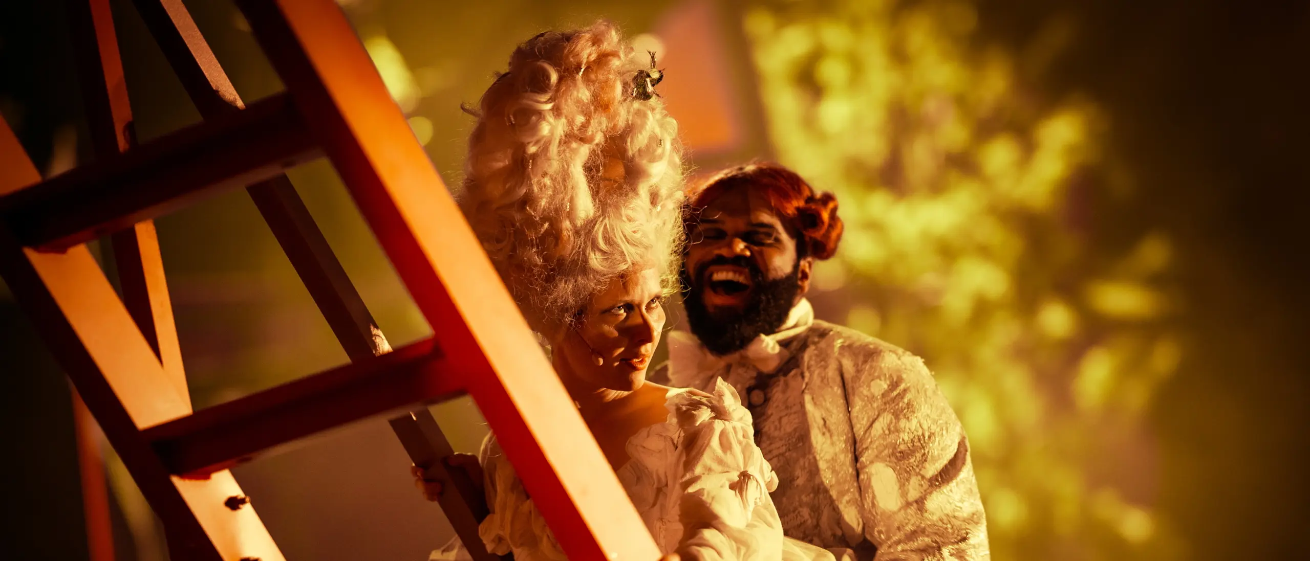 To skuespillere i overdådige, historiske kostymer smiler mens de står ved en rød stige i varmt scenelys.