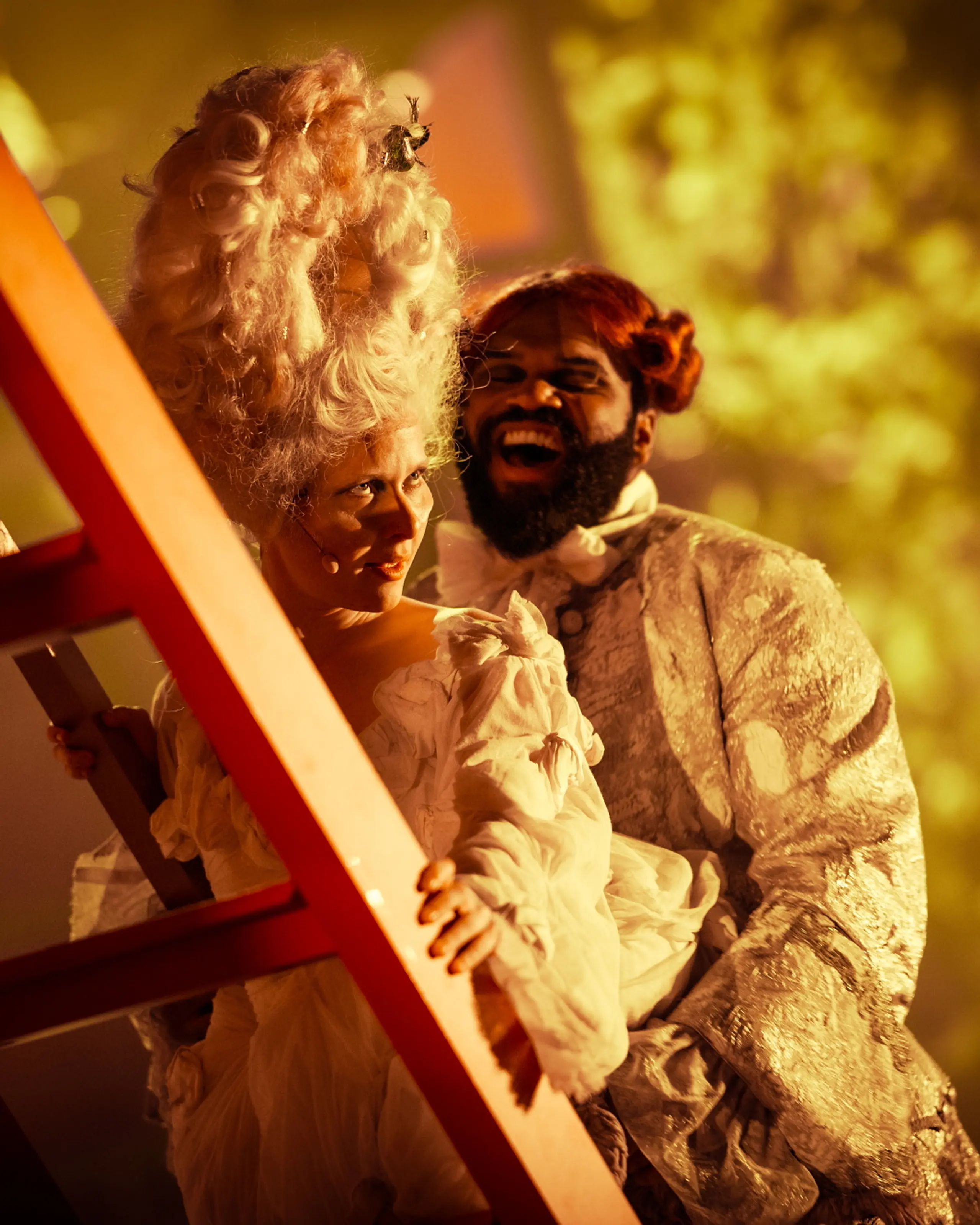 To skuespillere i overdådige, historiske kostymer smiler mens de står ved en rød stige i varmt scenelys.