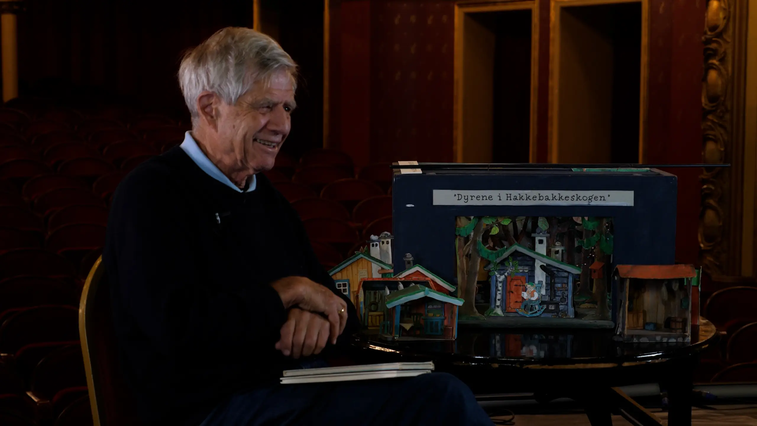 Eldre mann sitter ved siden av scenemodellen til Dyrene i Hakkebakkeskogen