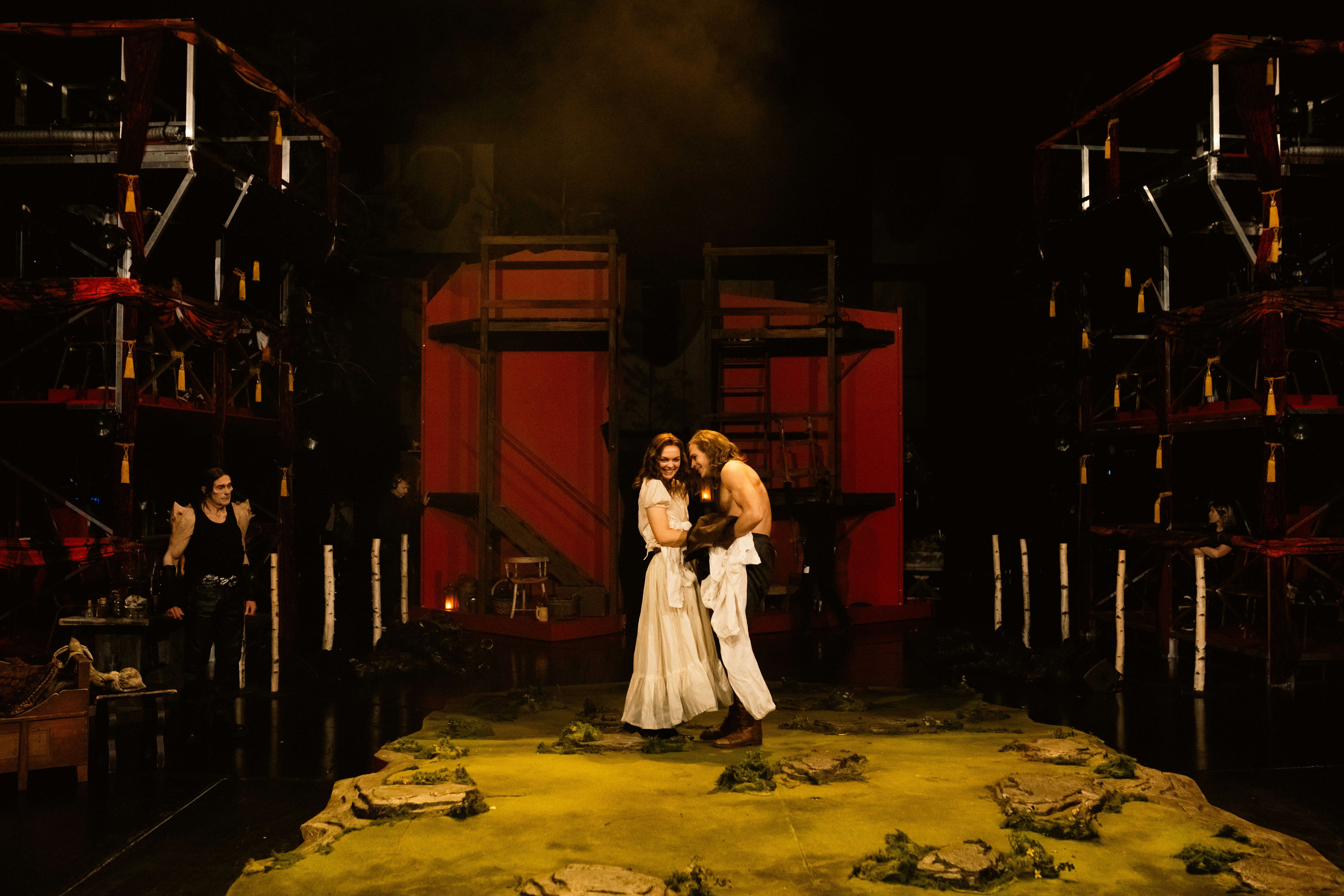 Scene med en kvinne og en skjorteløs mann som smiler til hverandre midt på scenen, omgitt av rød kulisse og mørklagte omgivelser.