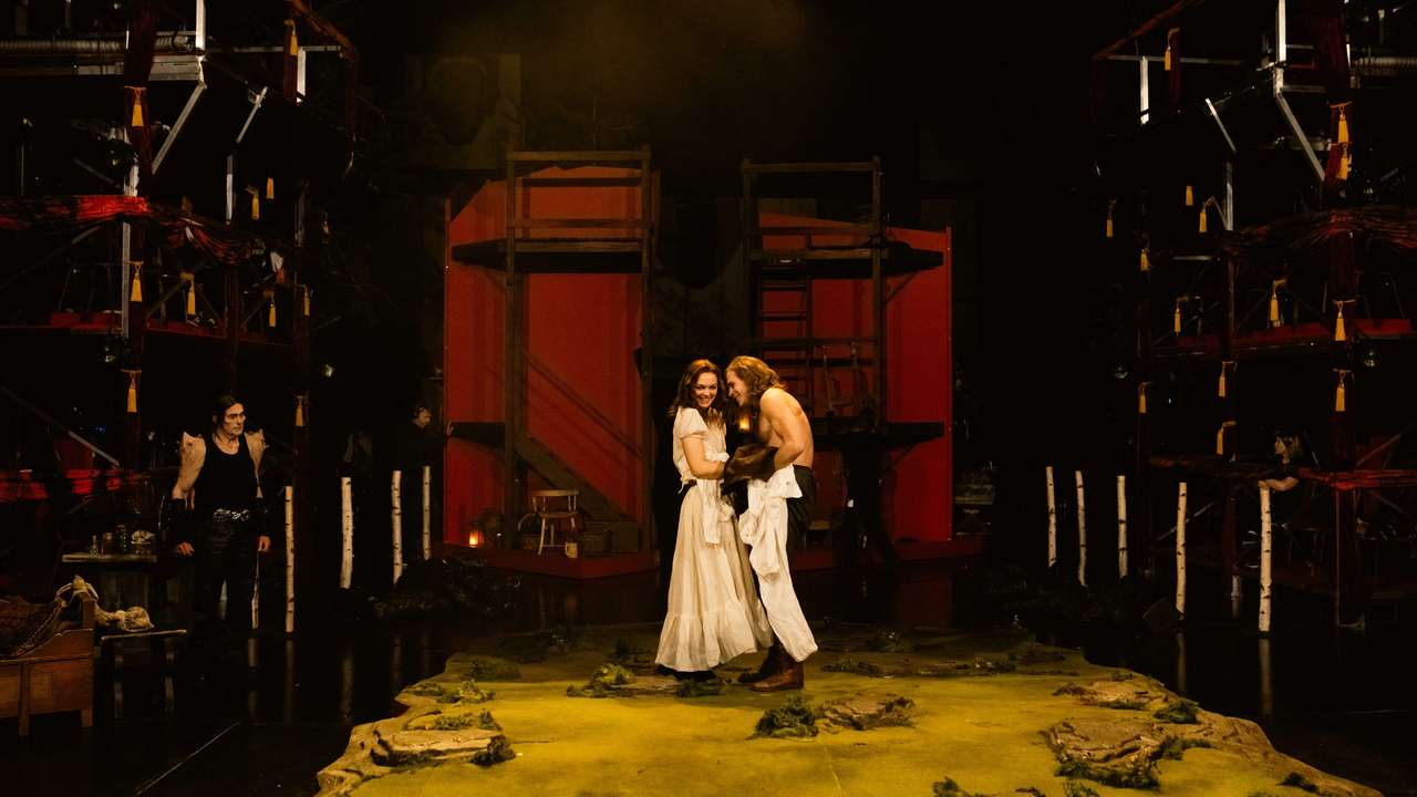 Scene med en kvinne og en skjorteløs mann som smiler til hverandre midt på scenen, omgitt av rød kulisse og mørklagte omgivelser.