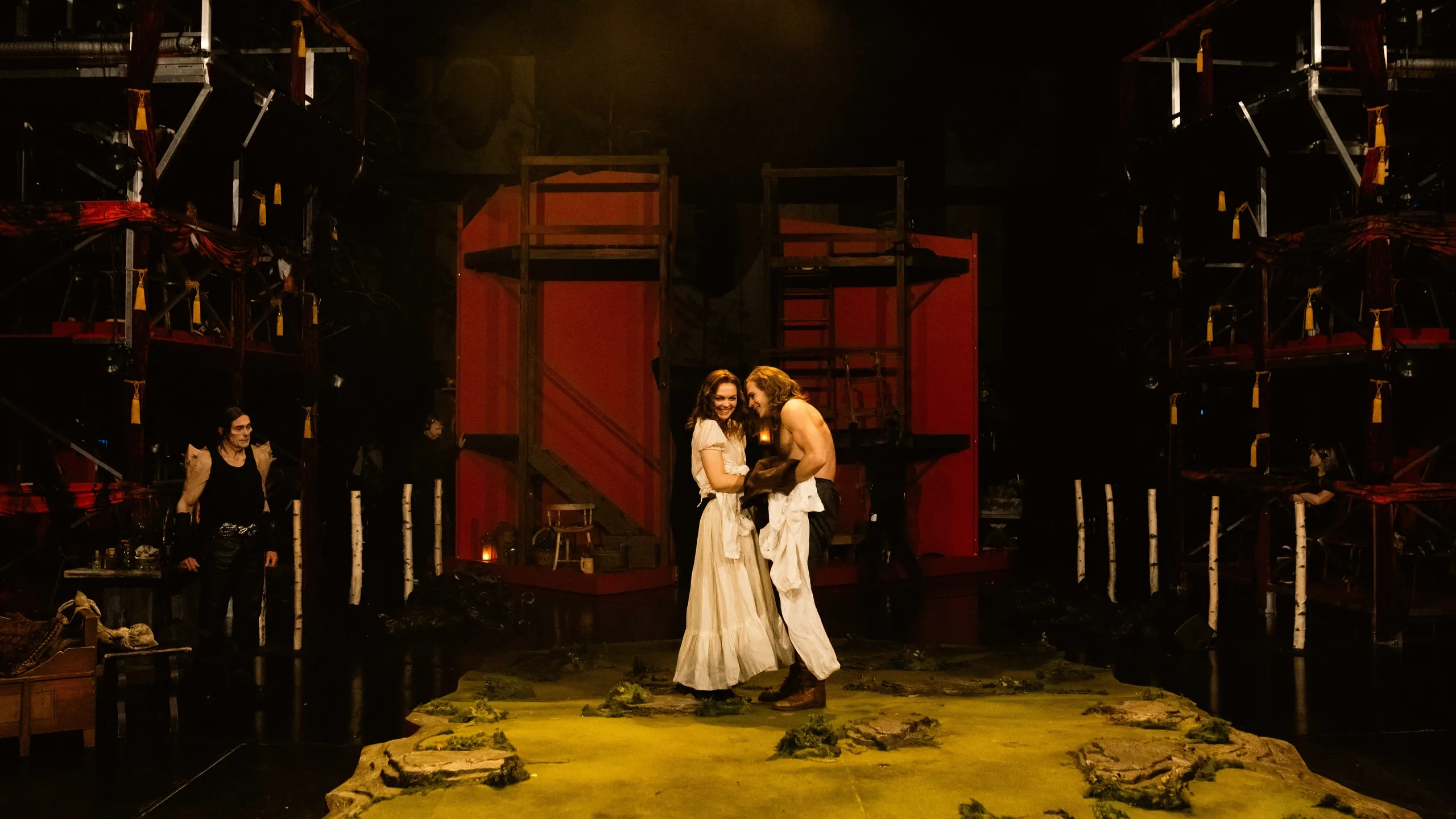 Scene med en kvinne og en skjorteløs mann som smiler til hverandre midt på scenen, omgitt av rød kulisse og mørklagte omgivelser.
