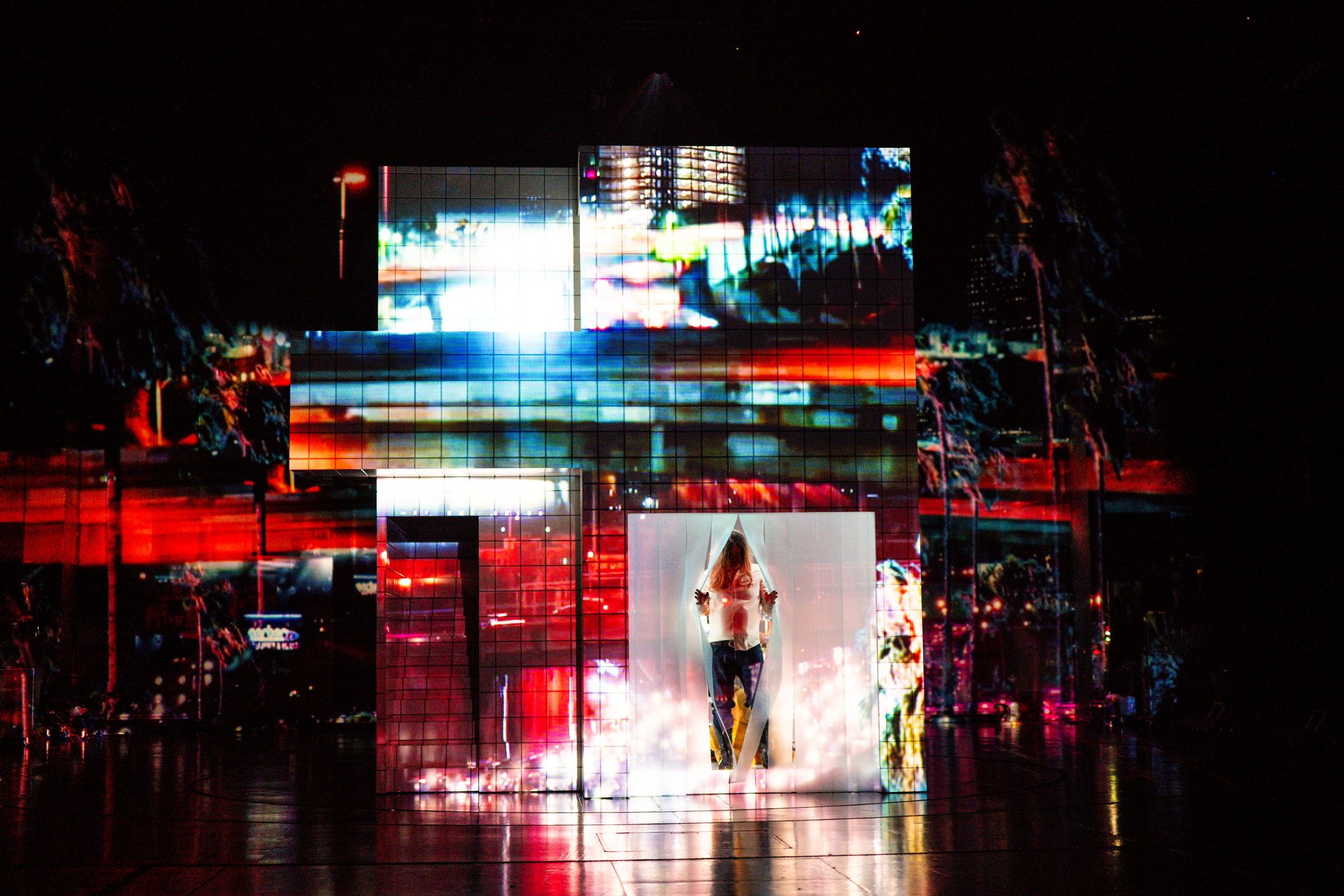 Skuespiller inne i transparent kube med projeksjoner av fargerike, urbane lys på fasadeformet struktur.